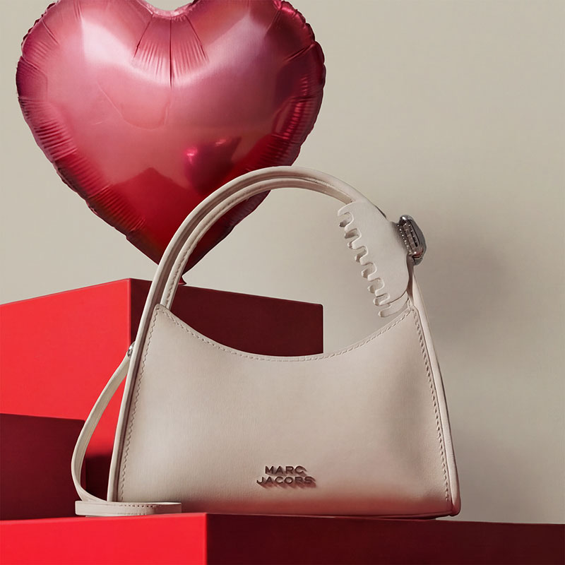 A beige handbag with a brand logo, placed next to a red heart-shaped balloon on red geometric blocks.