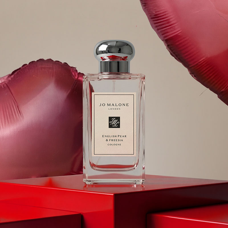 Perfume bottle of Jo Malone's English Pear & Freesia Cologne on red surface with heart-shaped balloons.