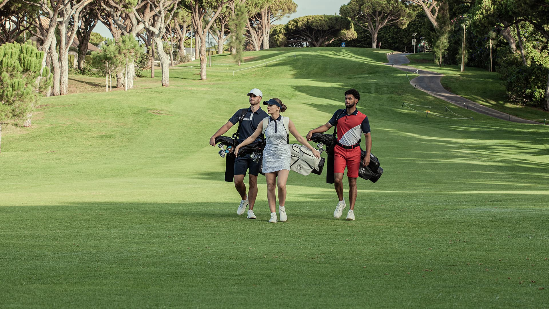 Three golfers walking across a golf course carrying their bags, wearing coordinated sport attire including polo shirts, caps and trainers.