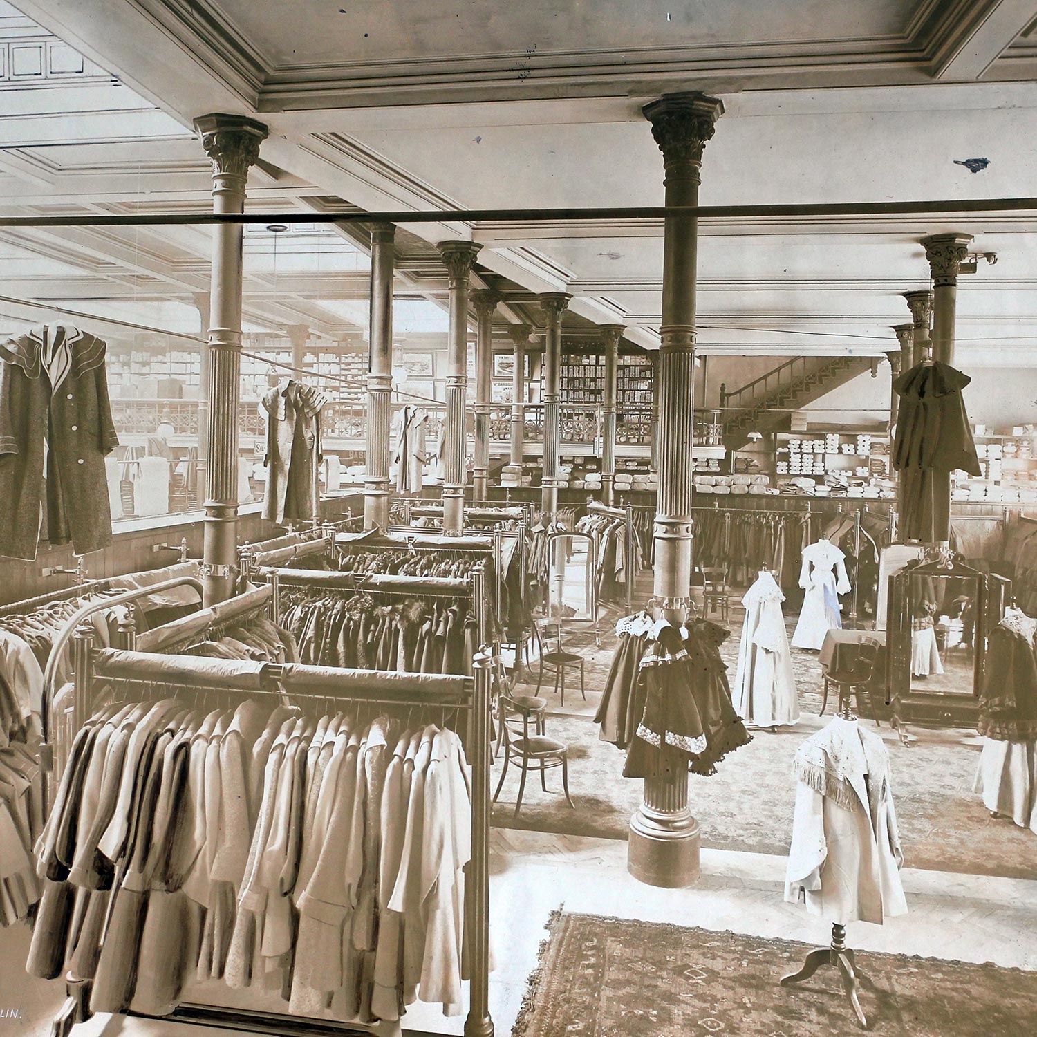 Interior of vintage department store with ornate columns, clothing rails displaying garments and mannequins dressed in period attire
