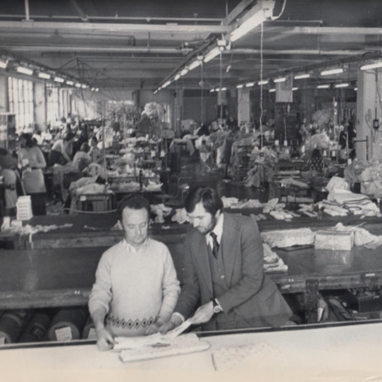 Black and white photograph of a clothing manufacturing workroom with numerous workers at sewing stations under fluorescent lighting