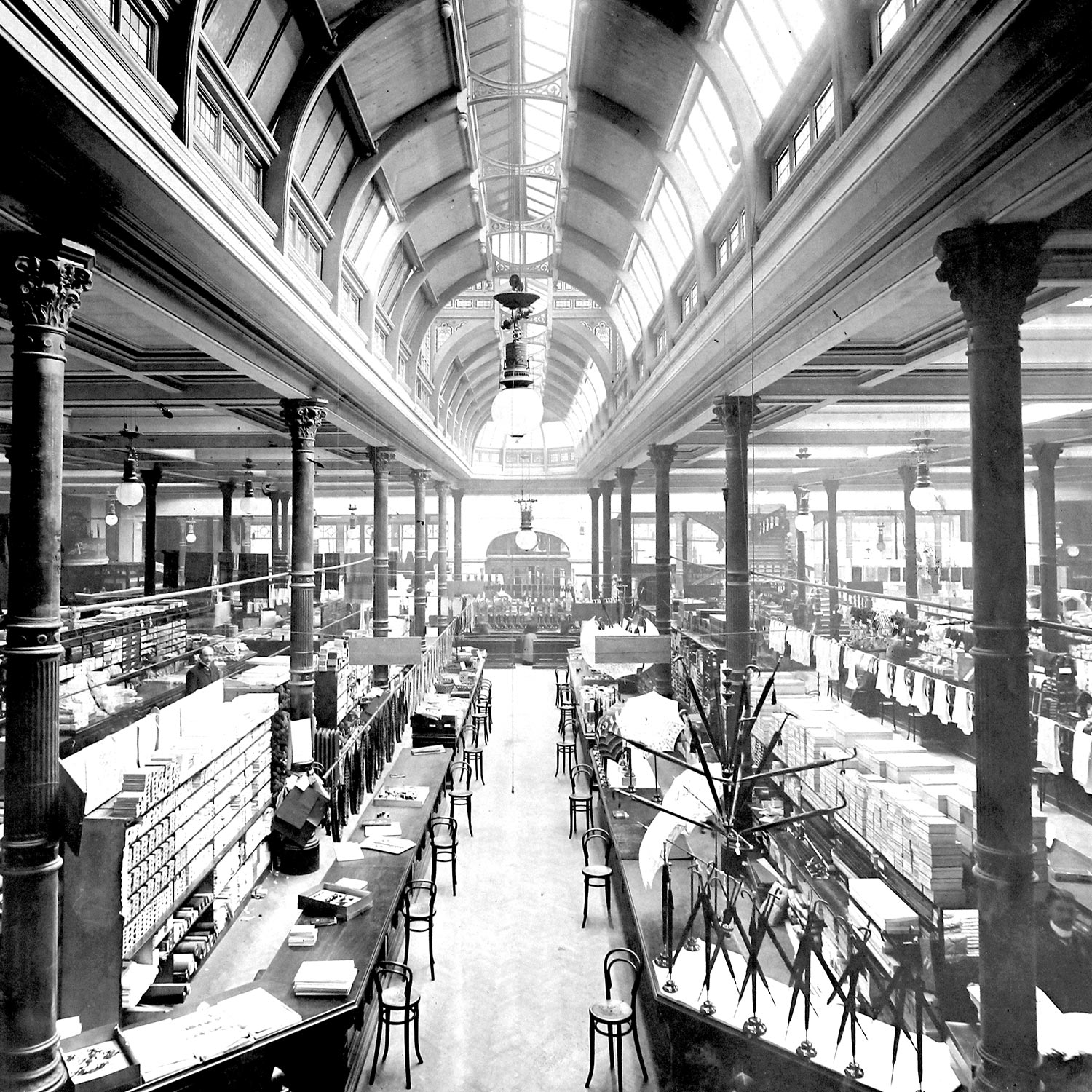 Historic black and white interior of a grand Victorian arcade with glass roof panels, ornate columns, and multiple levels of shop counters