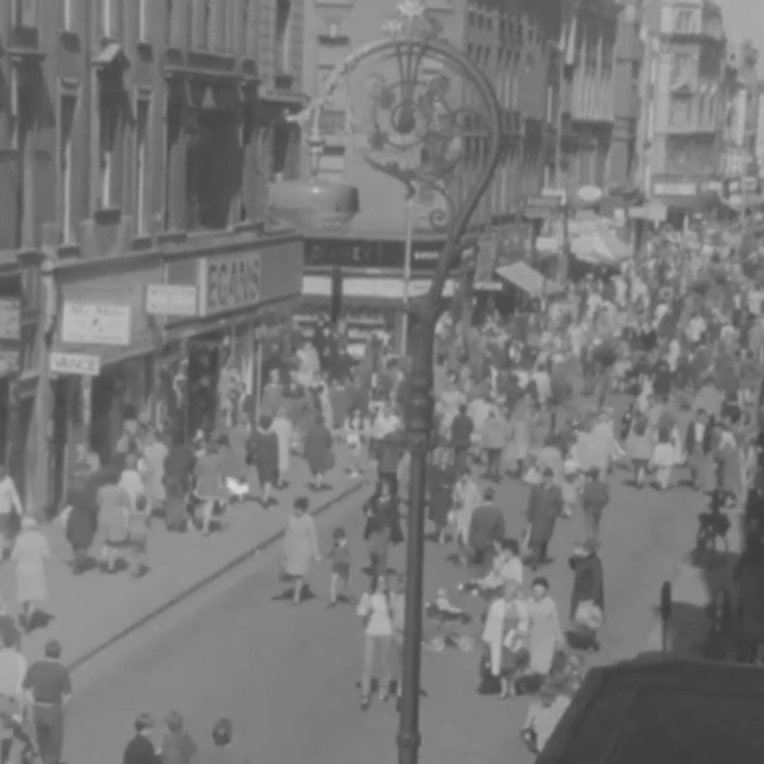 Black and white photograph of a busy pedestrianised street with crowds of shoppers, historic street lamp, and shop facades on Henry Street.
