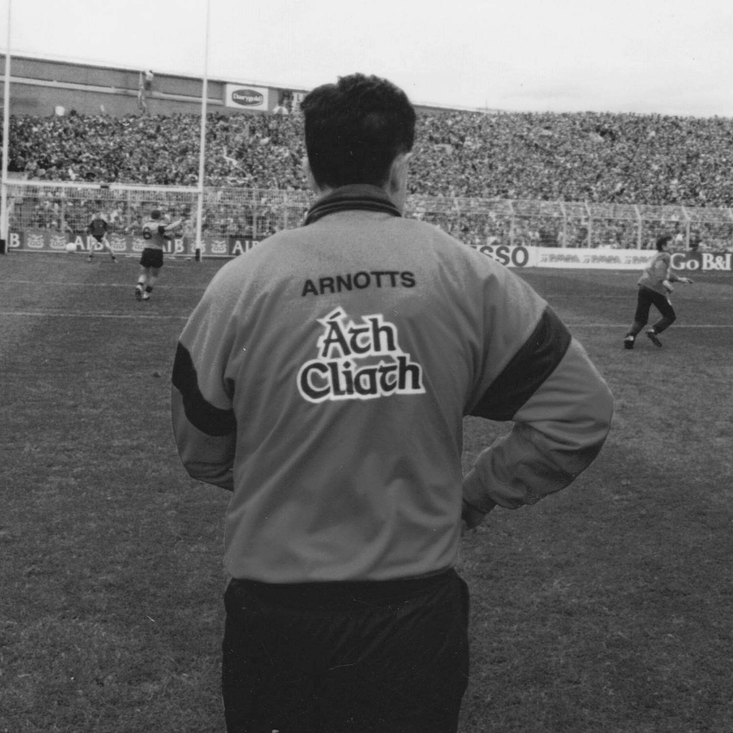 Black and white photograph of a man wearing an Arnotts Ach Cliath sports jacket at a crowded football stadium, viewed from behind.