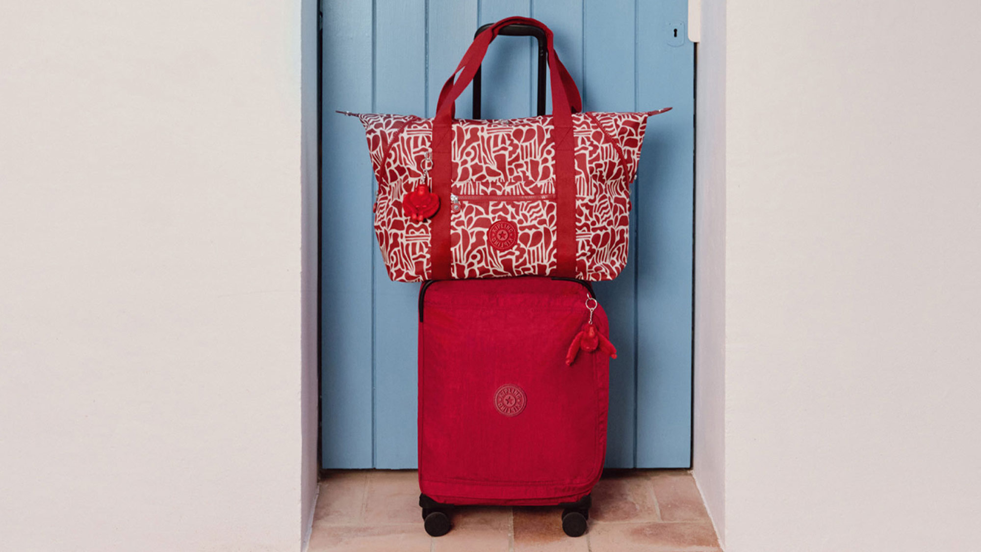 Red patterned tote bag and red cabin suitcase displayed against a blue door, promoting luggage and travel essentials