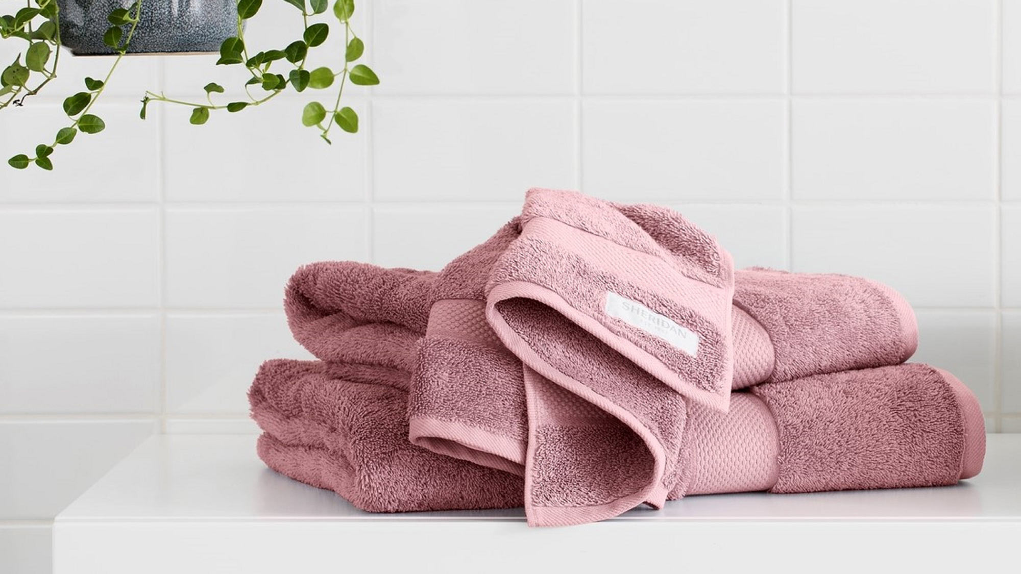 Pink towels stacked on a white counter with hanging plant and light bulb in a tiled bathroom.