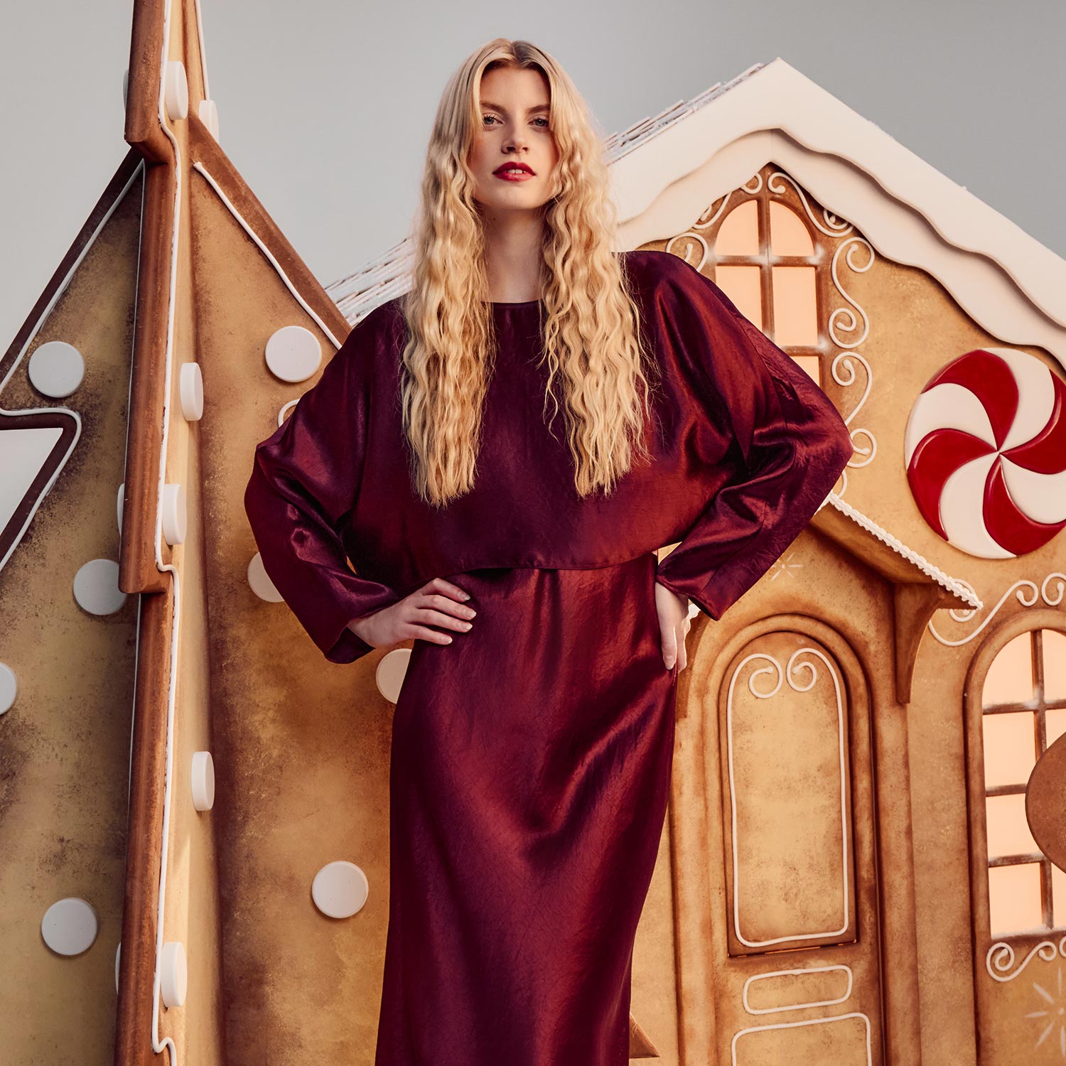 Woman in a maroon dress stands by gingerbread-themed decorations, including a house and a large cookie figure.