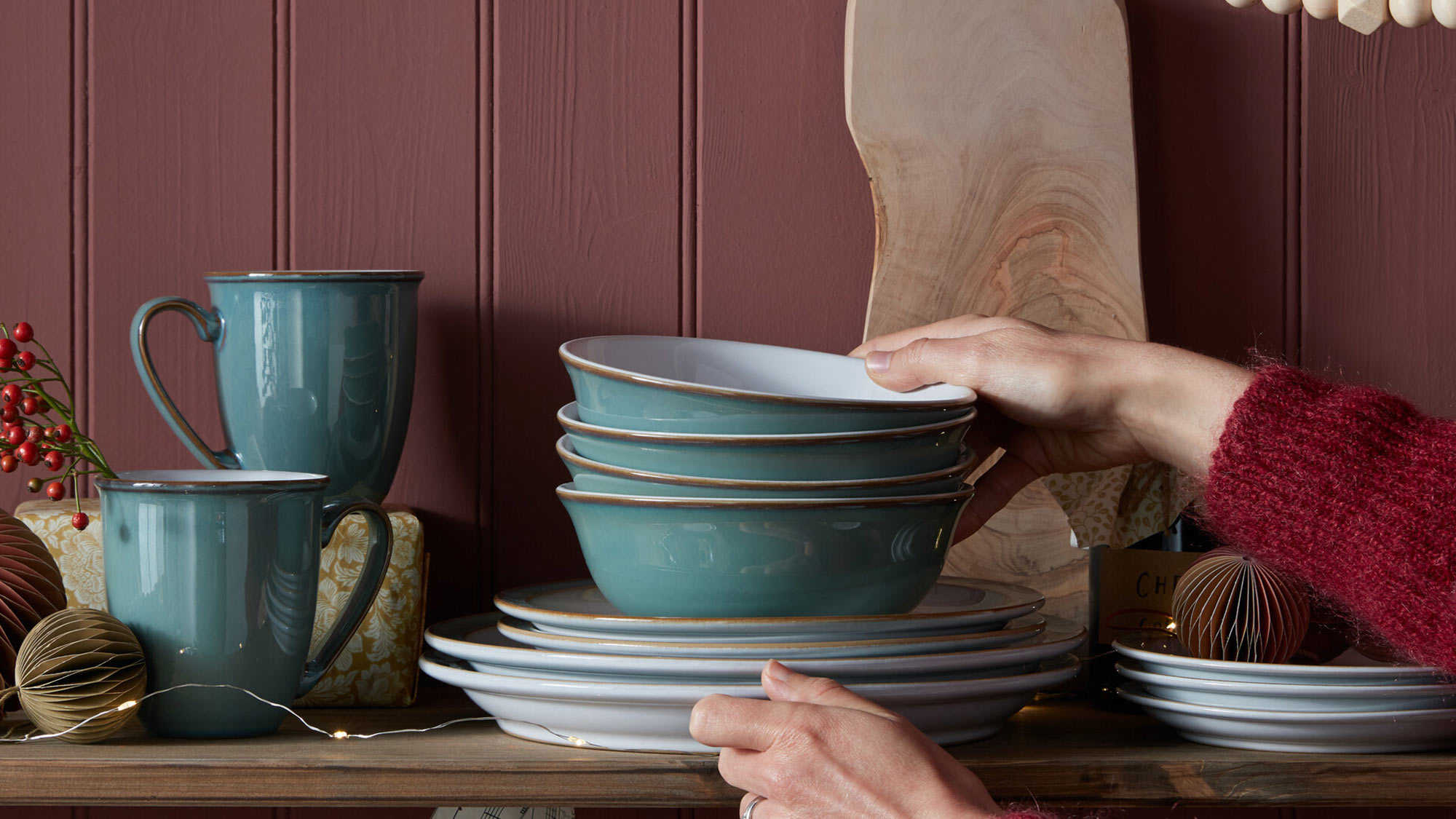 A person in a red sweater arranges teal dishes on a shelf with festive decor against a maroon background.