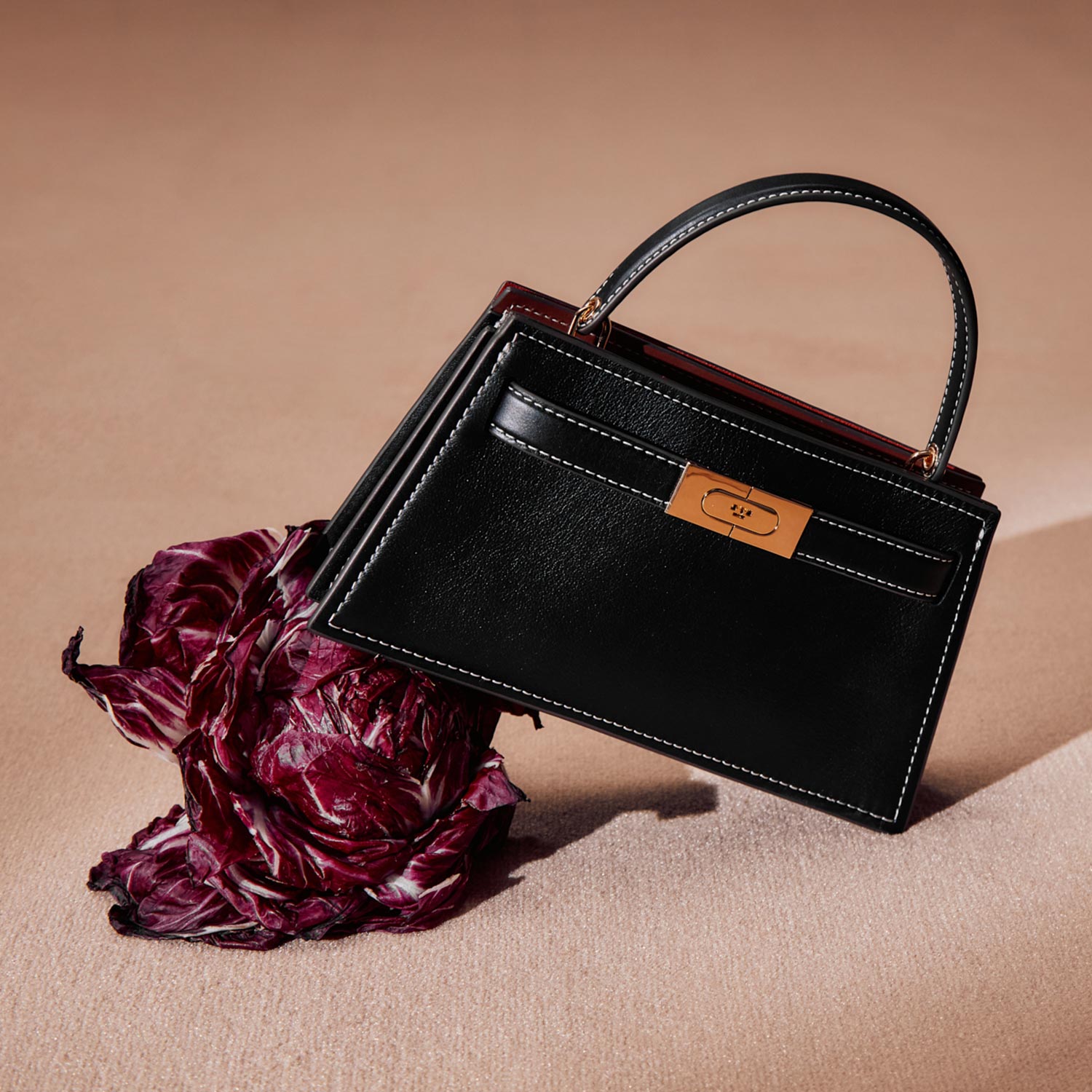 A black handbag with a handle, featuring a small tan tag, rests beside a piece of purple lettuce on a light pink surface.