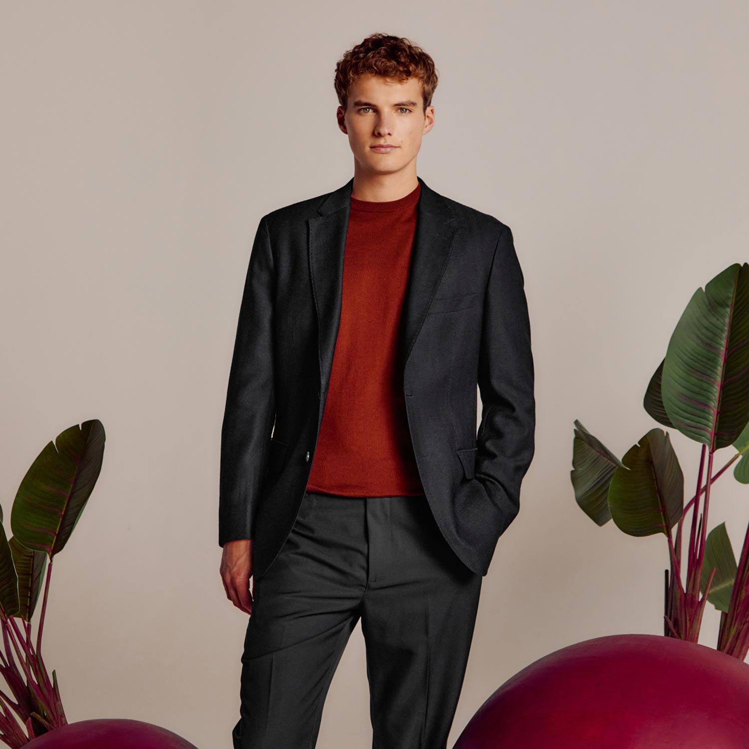 A person in a dark suit and red sweater stands between large purple spheres and green plants on a neutral background.