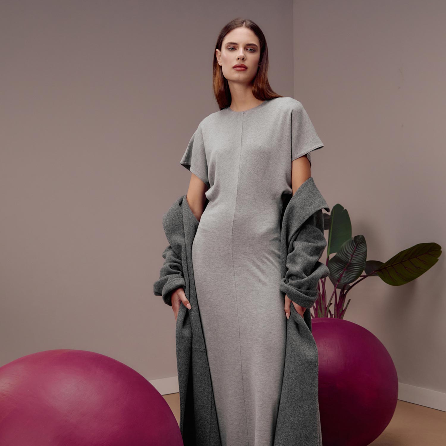 A woman in a gray dress and coat stands in a minimalist room with a large pink orb and a leafy plant.