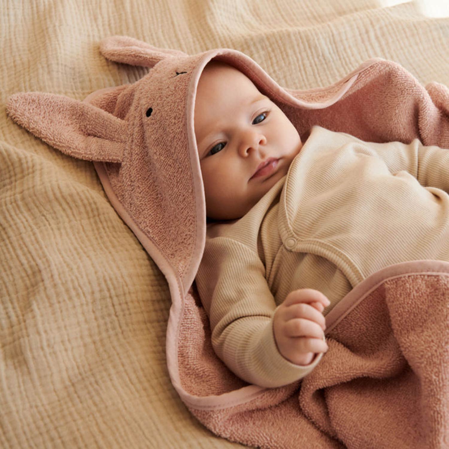 Newborn baby wrapped in brown bunny-eared hooded towel and cream romper on beige textured fabric