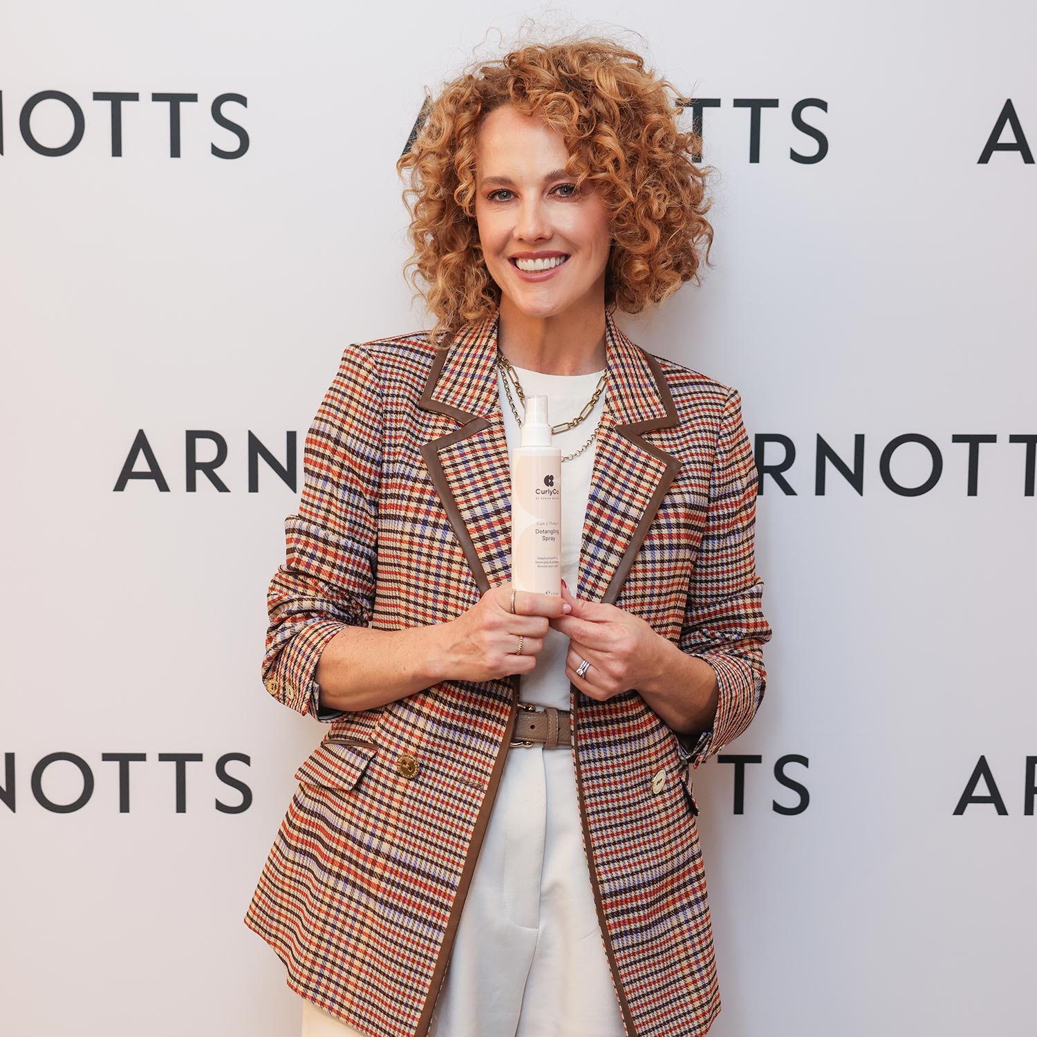 A person with curly hair holds a product in front of an Arnotts backdrop, wearing a plaid blazer and white pants.