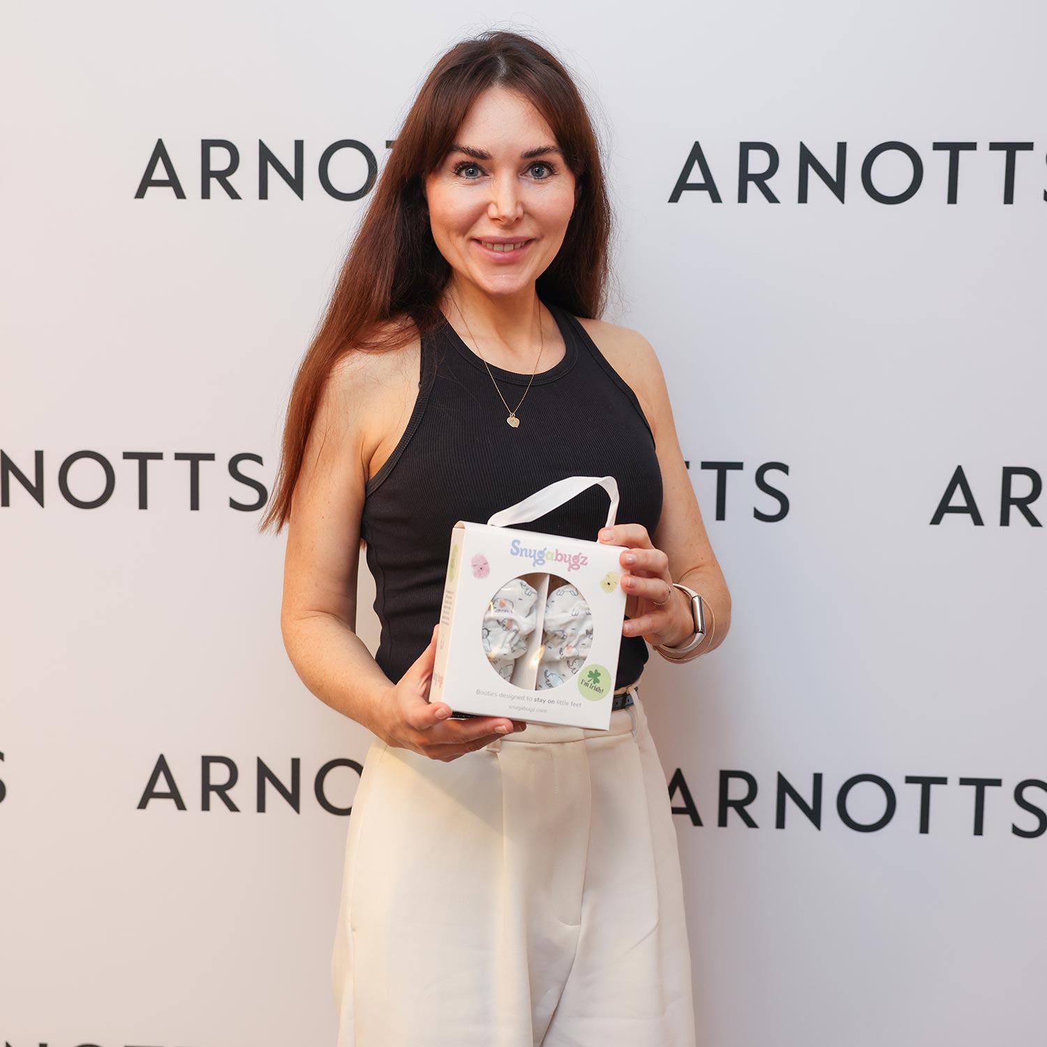 A woman in a black top and white pants holds a product box in front of an 'Arnotts' backdrop.