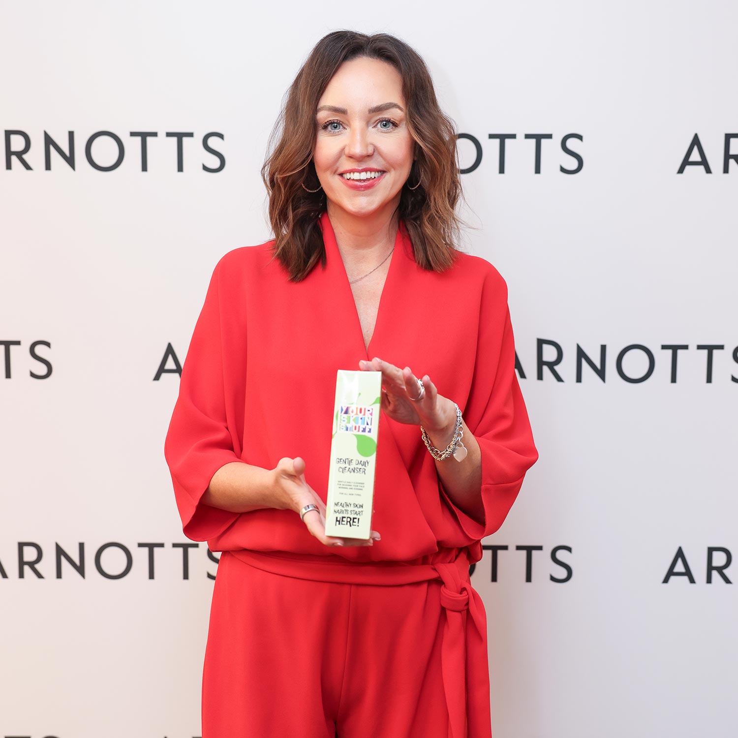 A woman in a red outfit holds a booklet, smiling in front of an 'ARNOTTS' backdrop.