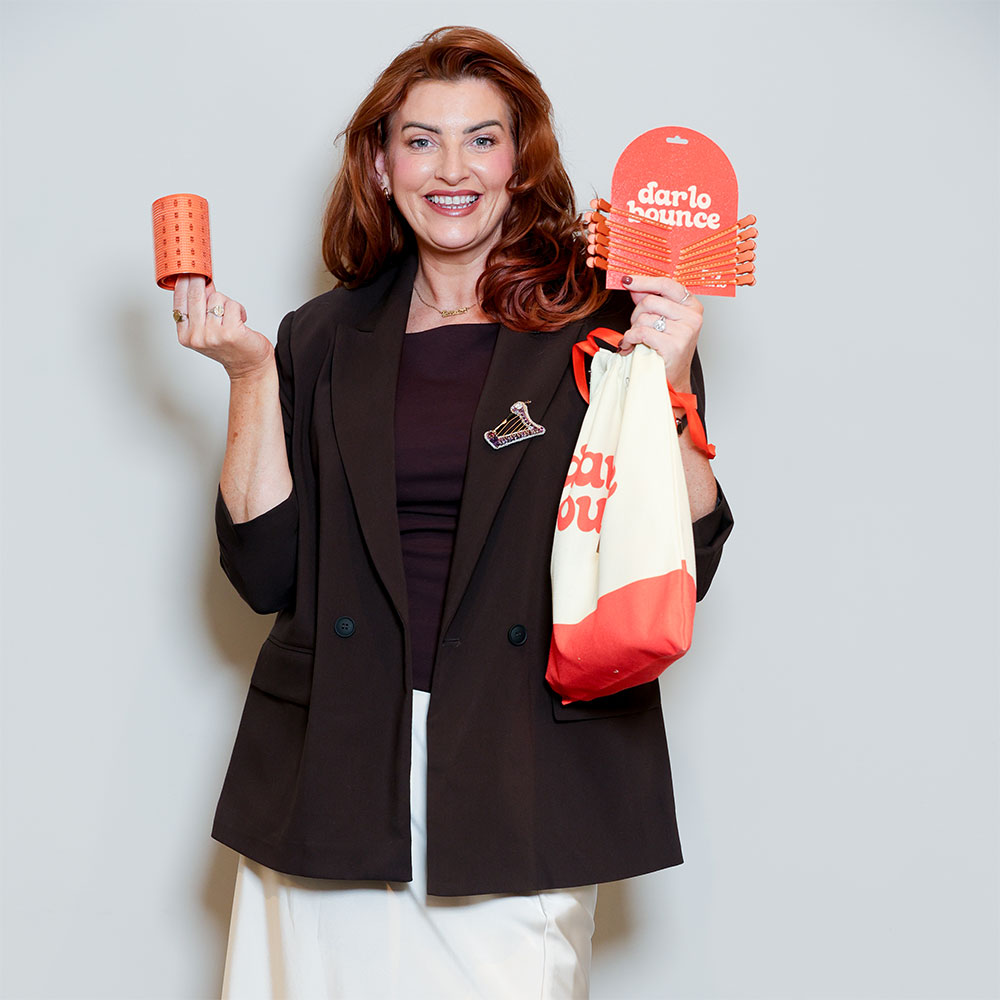 Woman holding orange Darlo Bounce hair rollers and product packaging, wearing black blazer