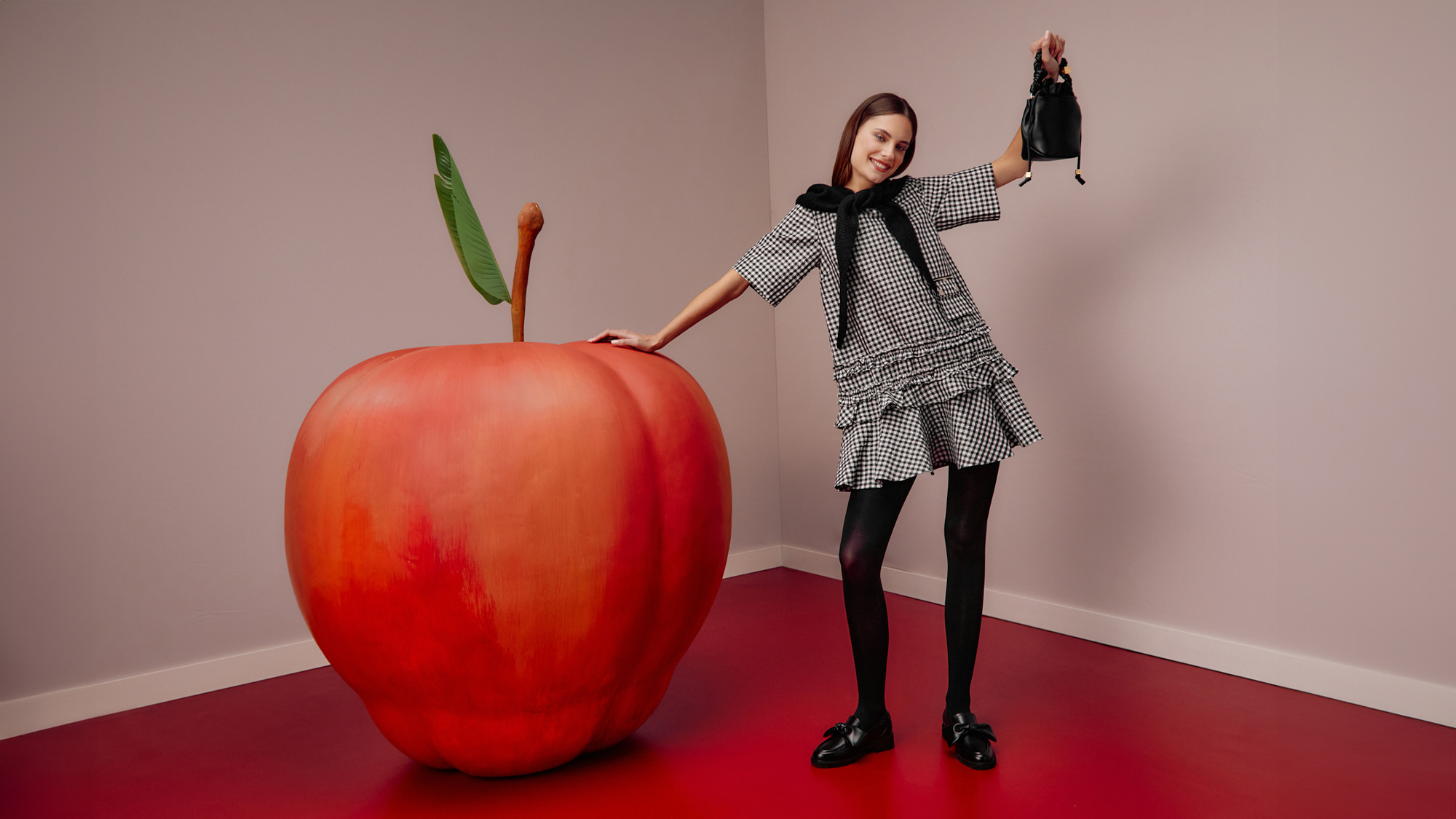 Person in a patterned dress poses with a giant apple, holding a small black bag, in a room with red flooring.