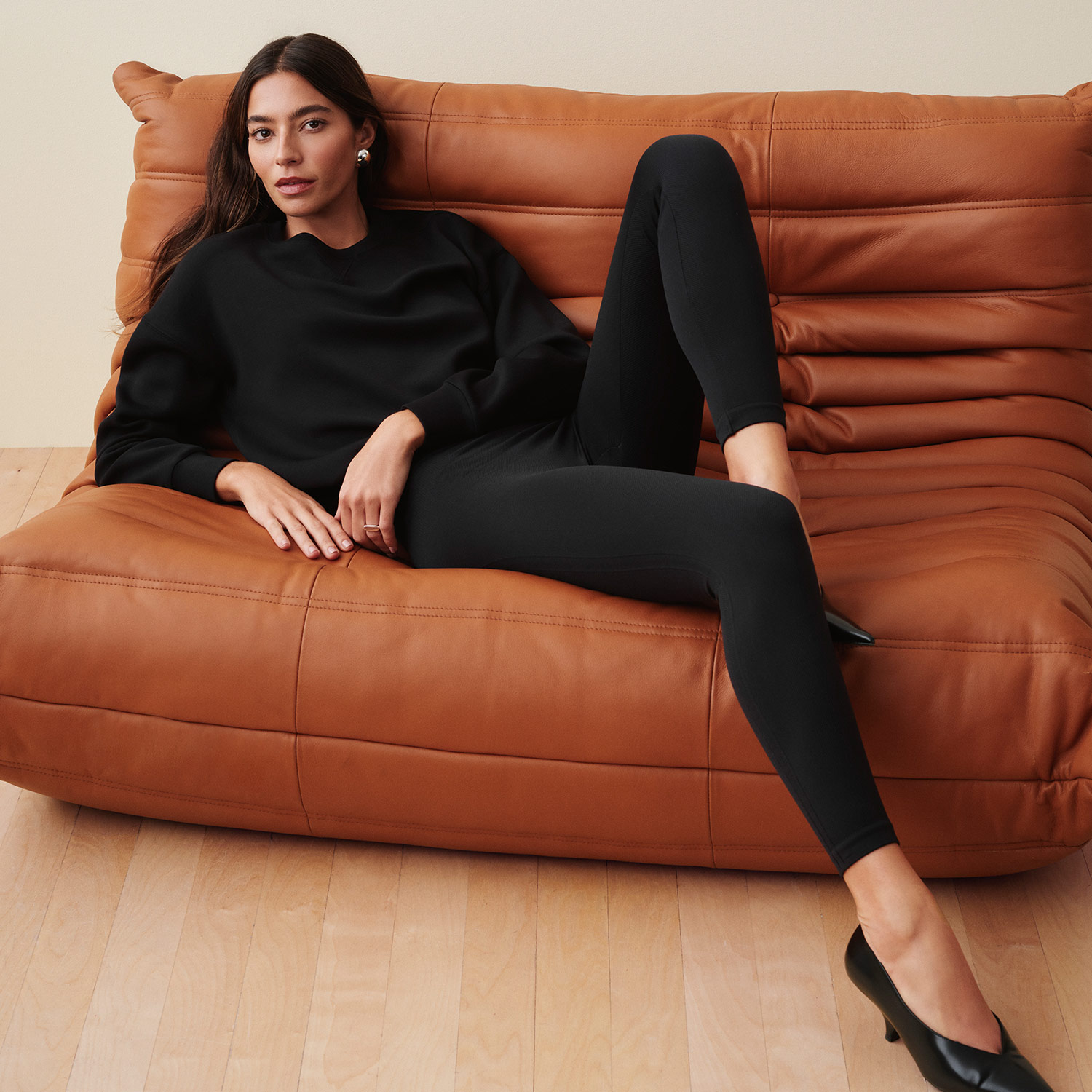 A woman in black clothing reclines on a brown sofa, wearing black heels.