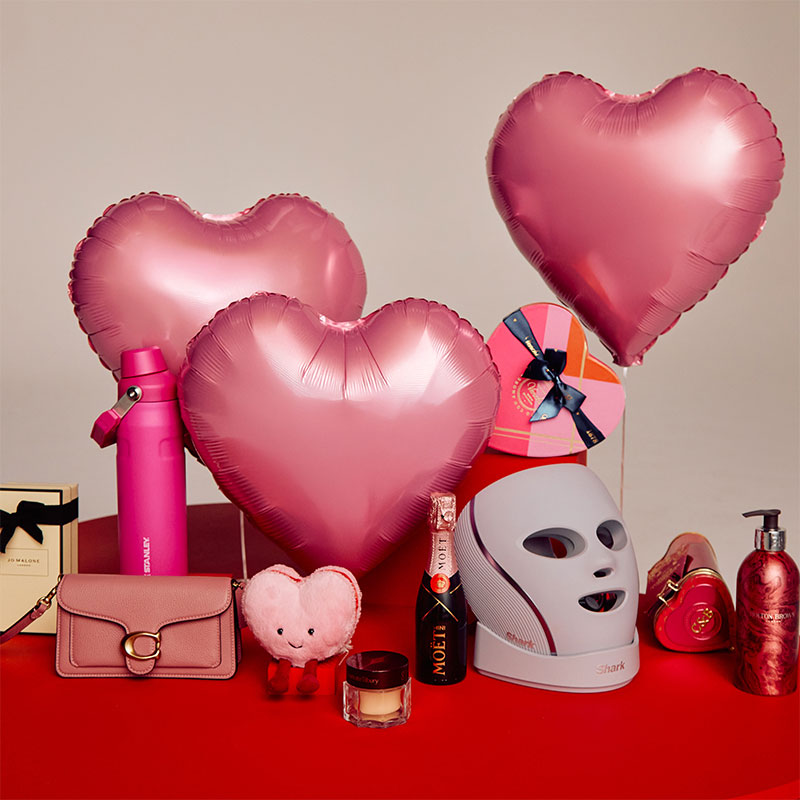 A collection of pink-themed items including heart-shaped balloons, a water bottle, skincare products, a purse, and a champagne bottle on a red surface.