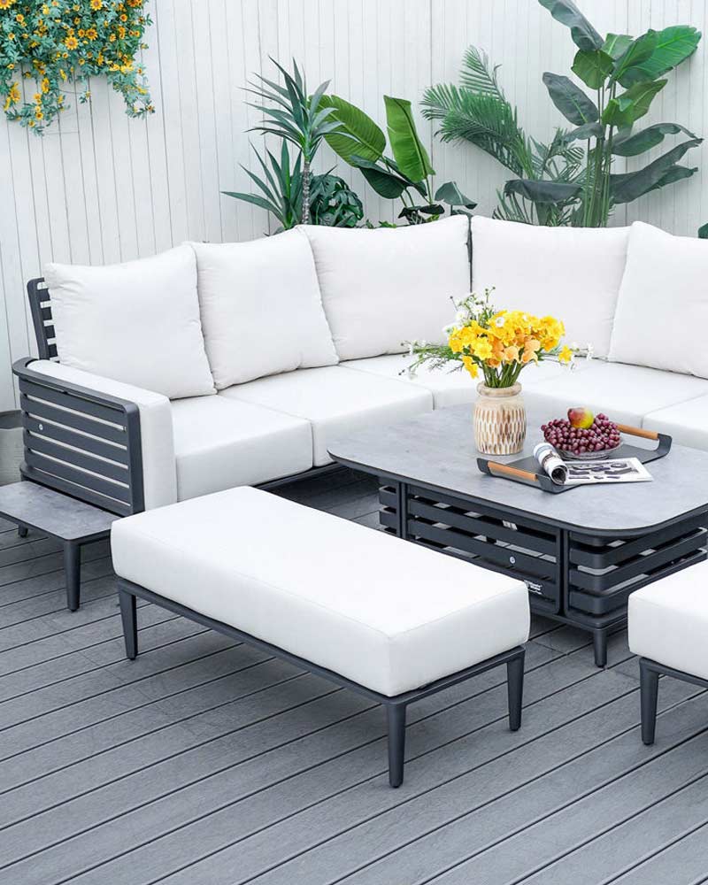 Outdoor patio with white cushioned furniture, a gray table featuring a vase of yellow flowers, fruit, and greenery on the wall.