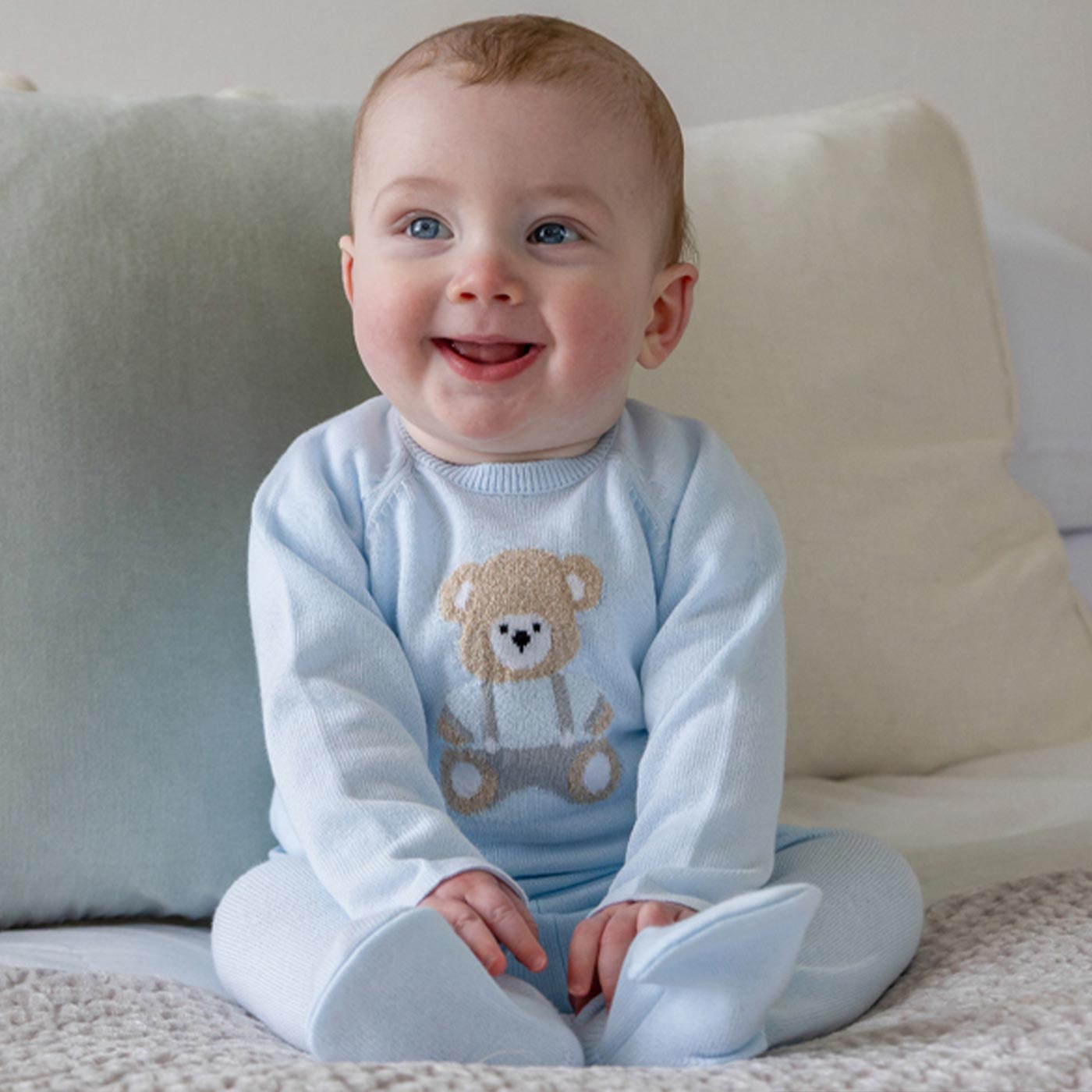 Smiling baby wearing light blue knit jumper with teddy bear design and white leggings, sitting on neutral cushions
