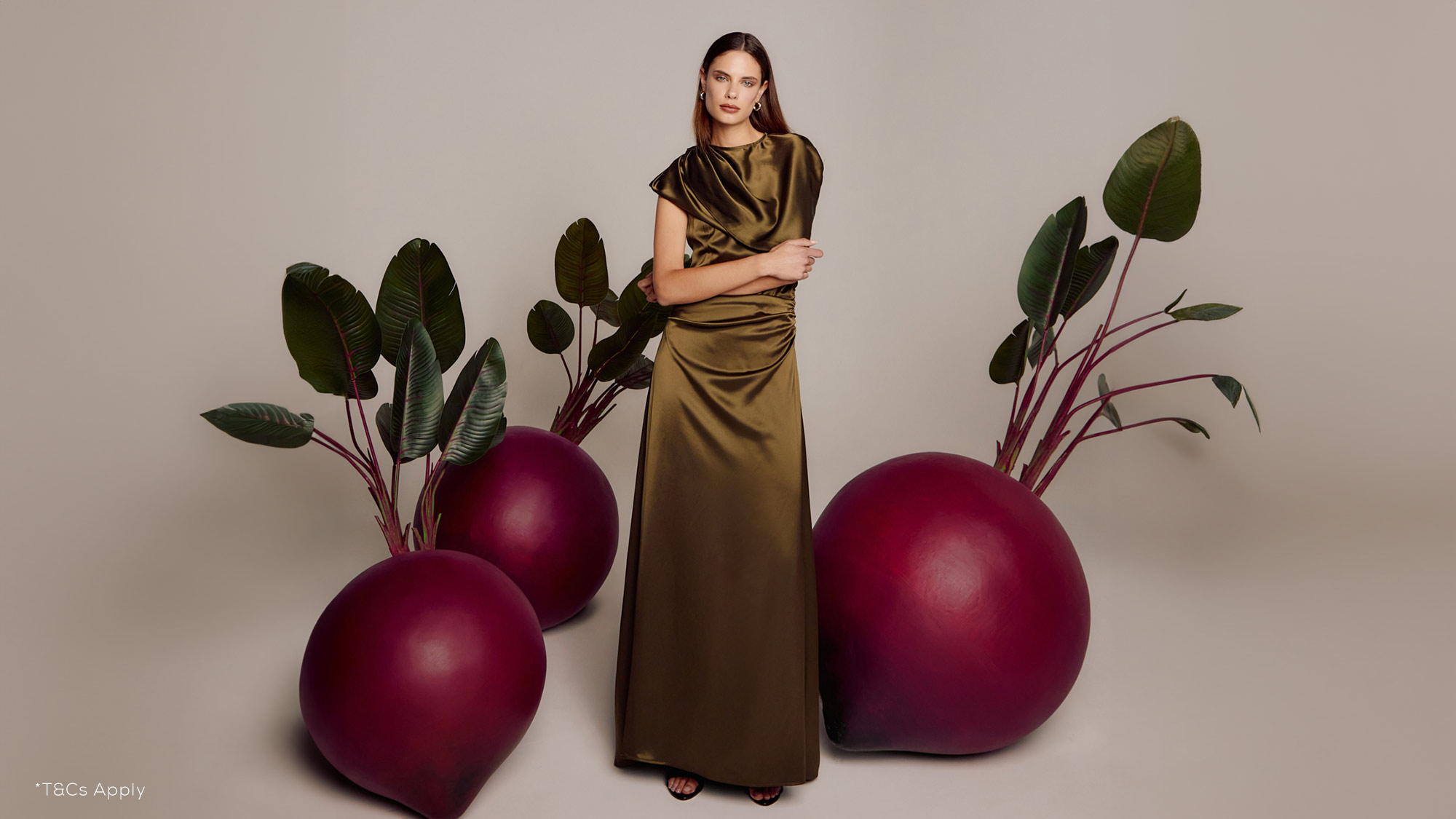 A woman in an olive dress stands among large, stylized beetroots with lush green leaves against a neutral background.