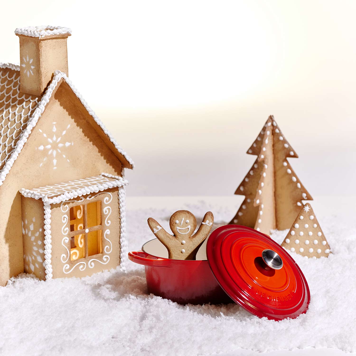 A gingerbread house and tree on snow with a gingerbread man in a red pot.
