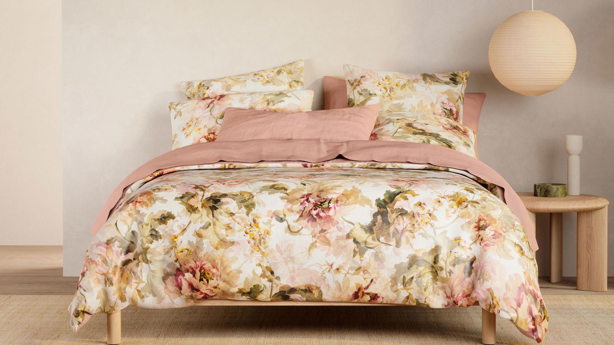 Cozy bedroom with floral bedding, pink accents, a round pendant lamp, and a small wooden side table with a candle.