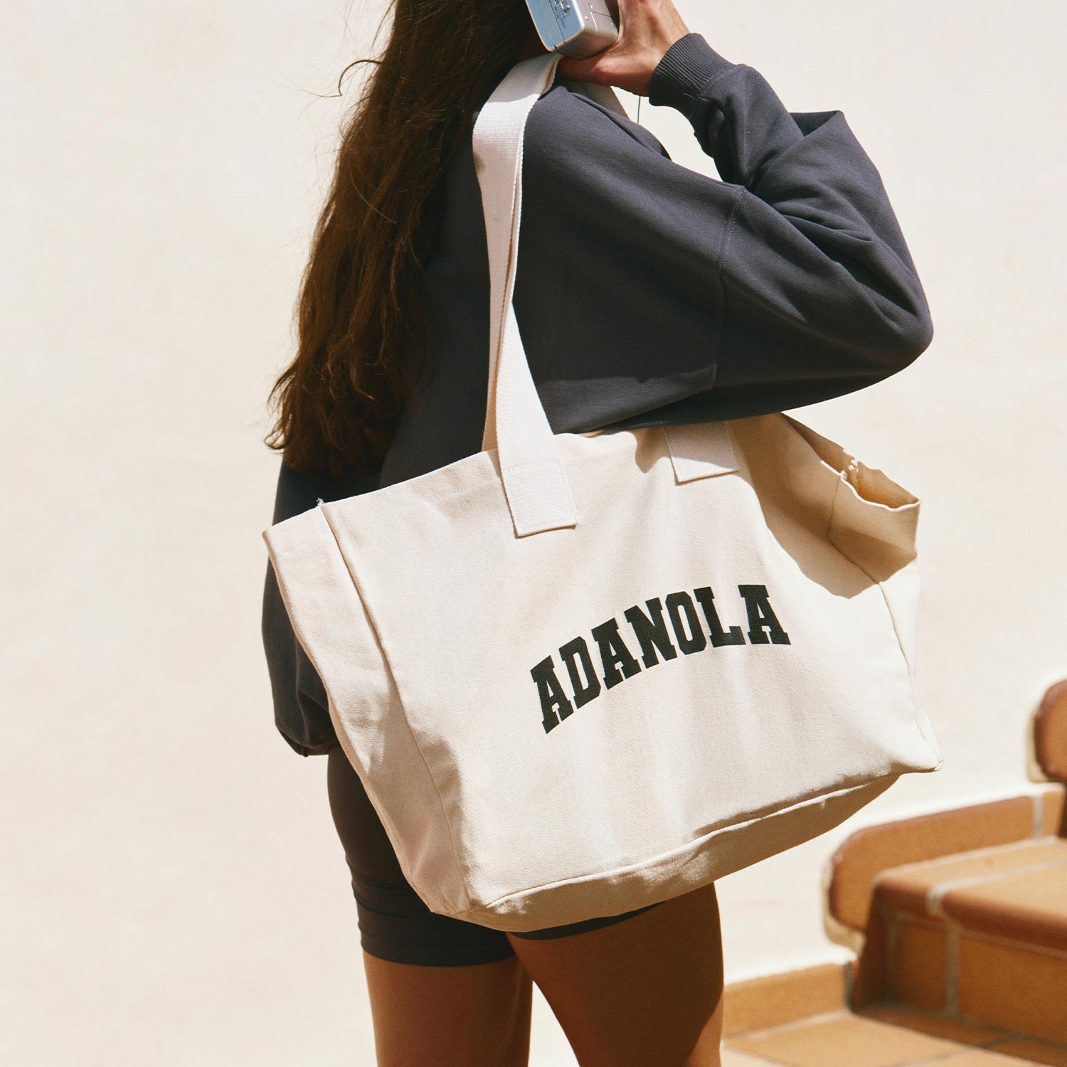 A woman walks with a large tote bag labeled 'ADANOLA,' wearing sneakers, white socks, and dark clothing, holding a phone.