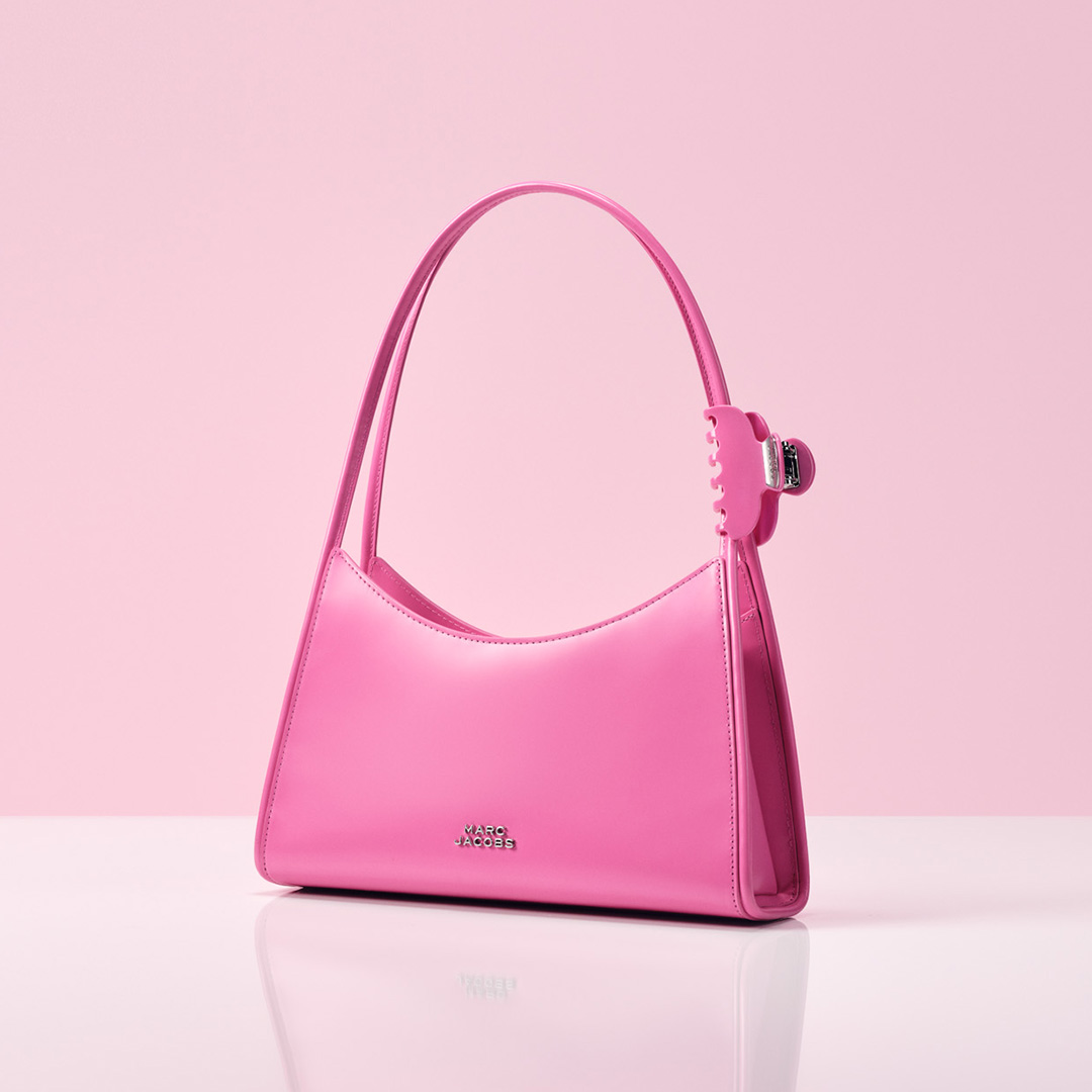 A pink handbag is placed on a reflective surface against a light pink background.