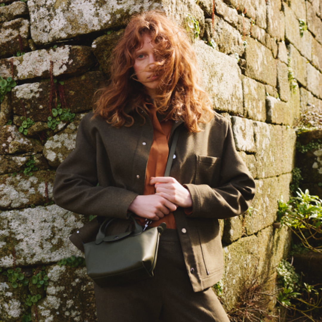 A person with wavy hair stands against a mossy stone wall, wearing a dark jacket and holding a small green bag.