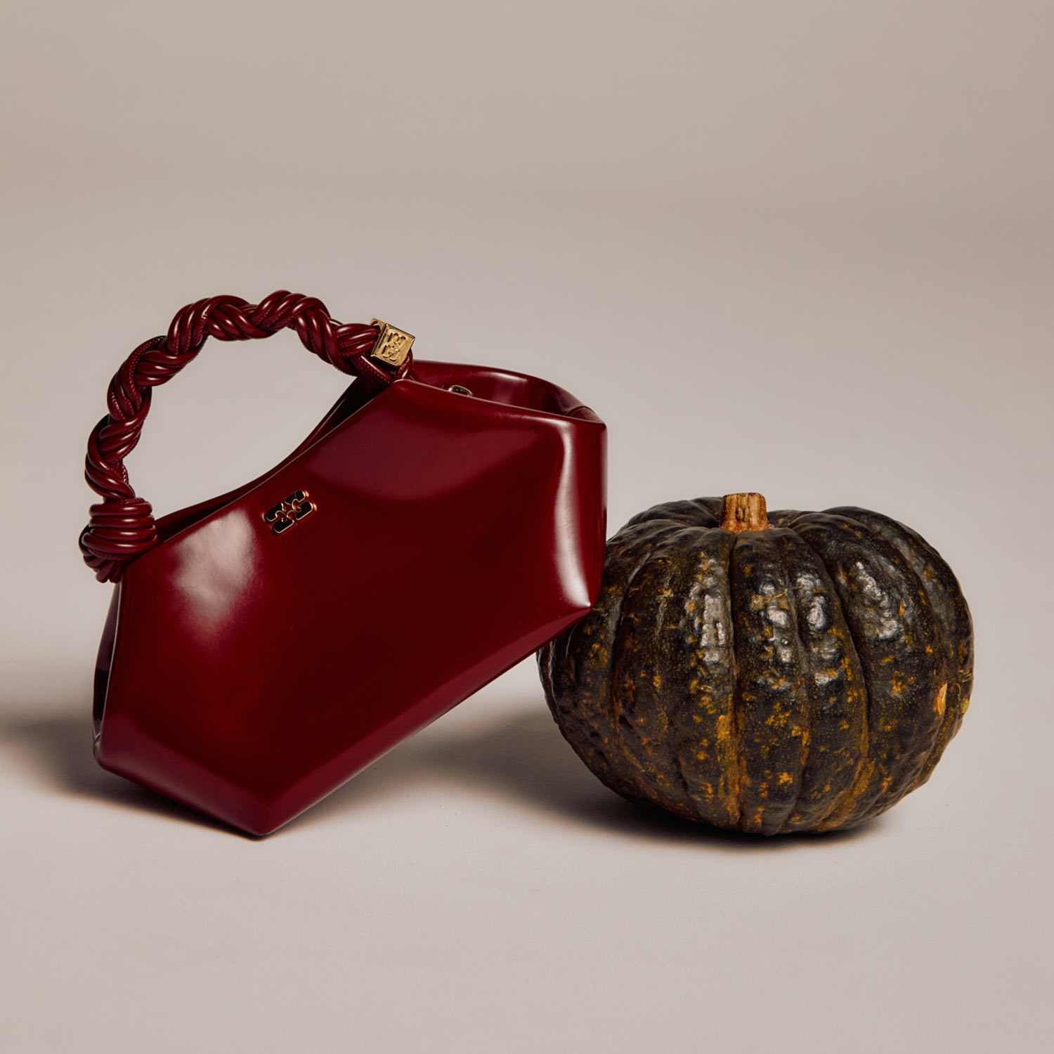 A dark red handbag with a twisted handle is placed next to a small, dark green pumpkin on a neutral background.