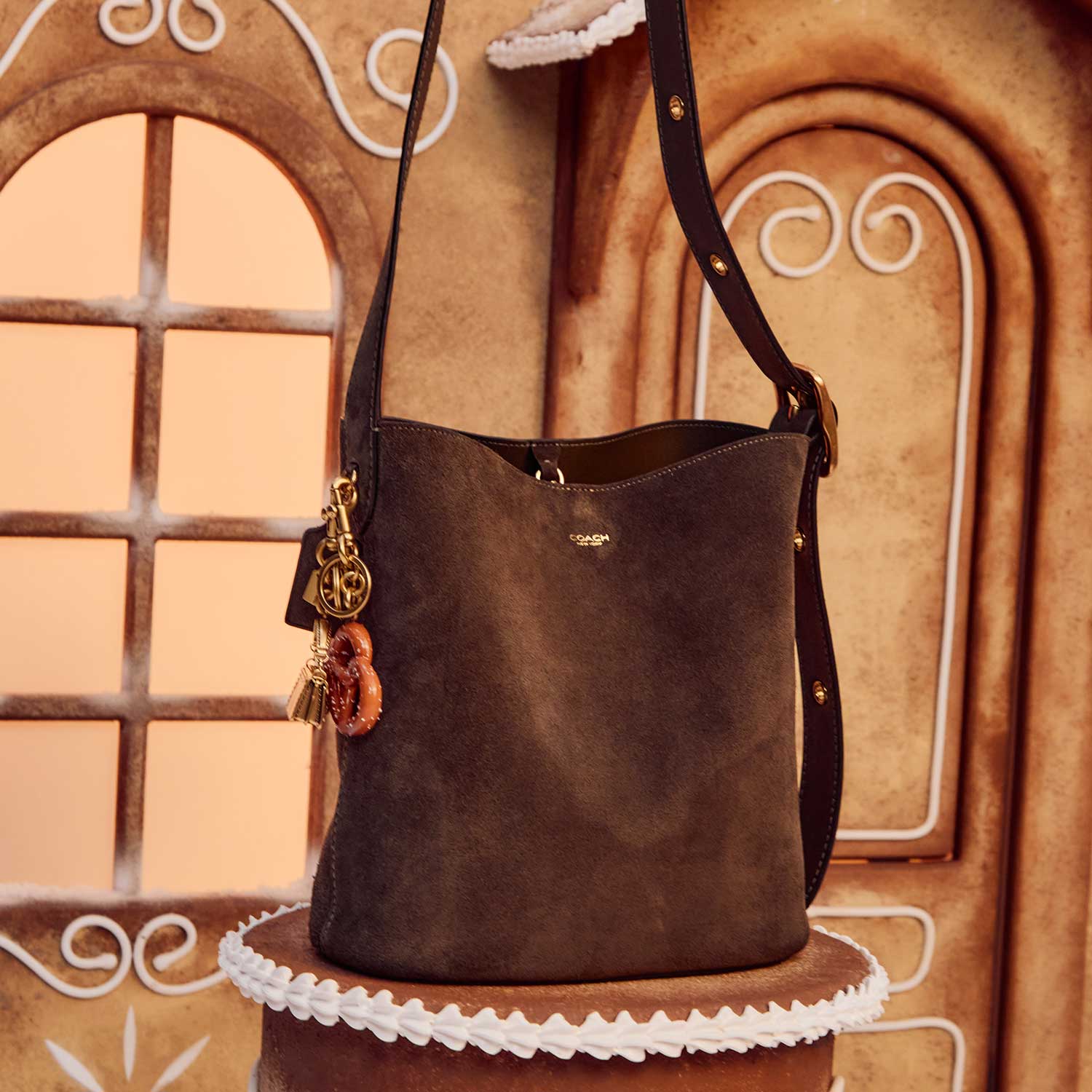 A brown handbag with a strap and charms is displayed against a gingerbread house background.