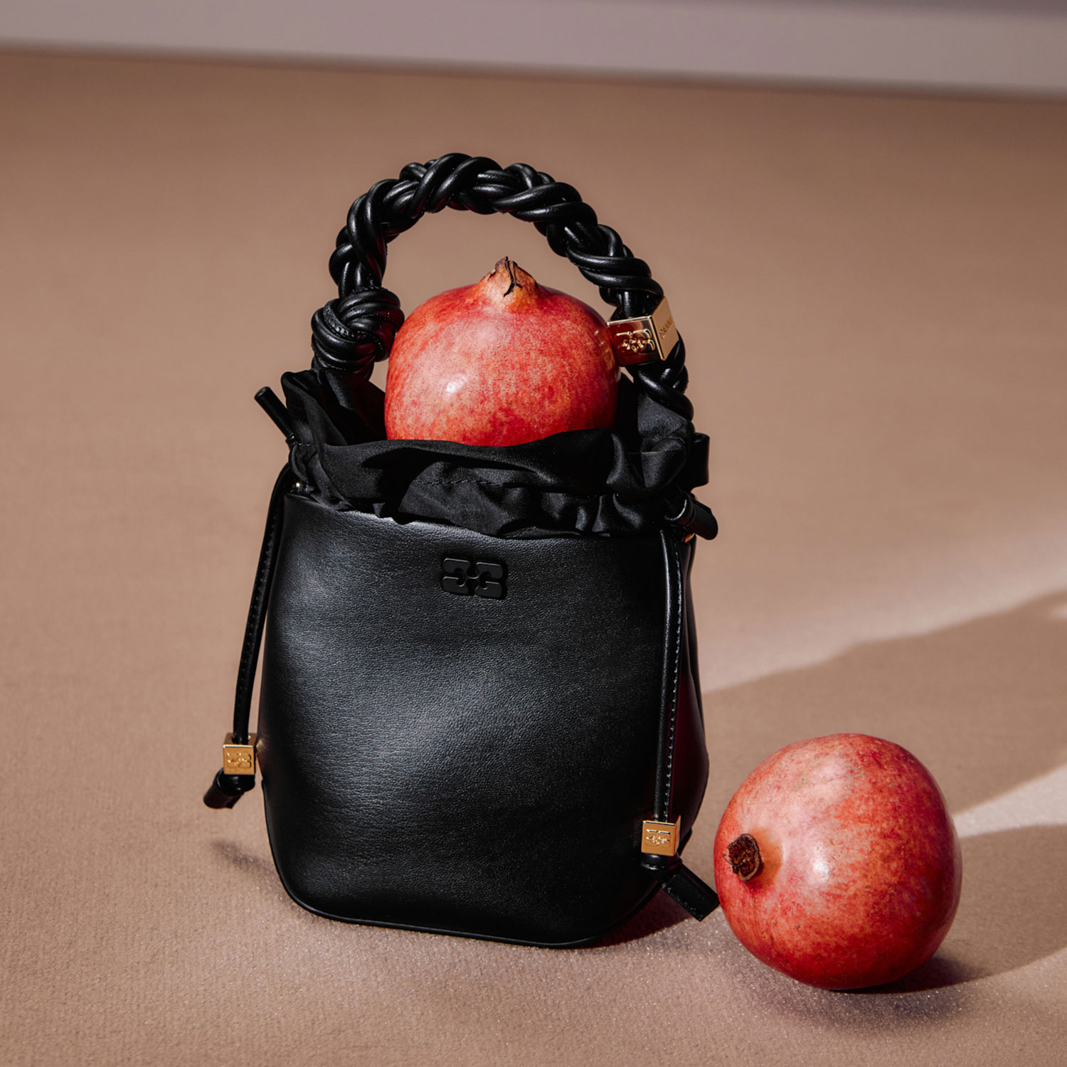 Black handbag with a braided handle, containing a pomegranate, with another pomegranate beside it on a neutral background.