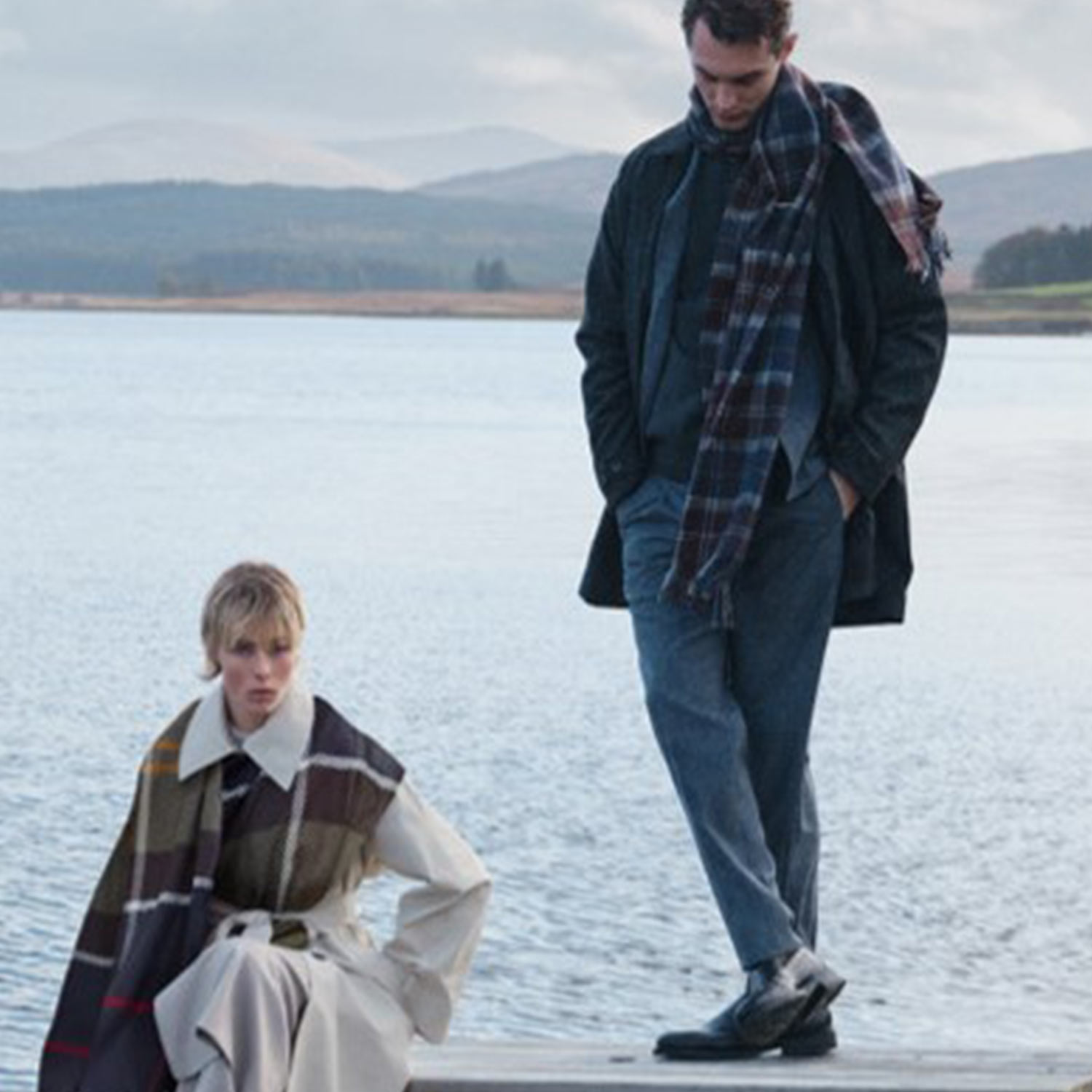 Two people in stylish clothing with scarves stand and sit on a dock by a calm lake, with hills in the background.
