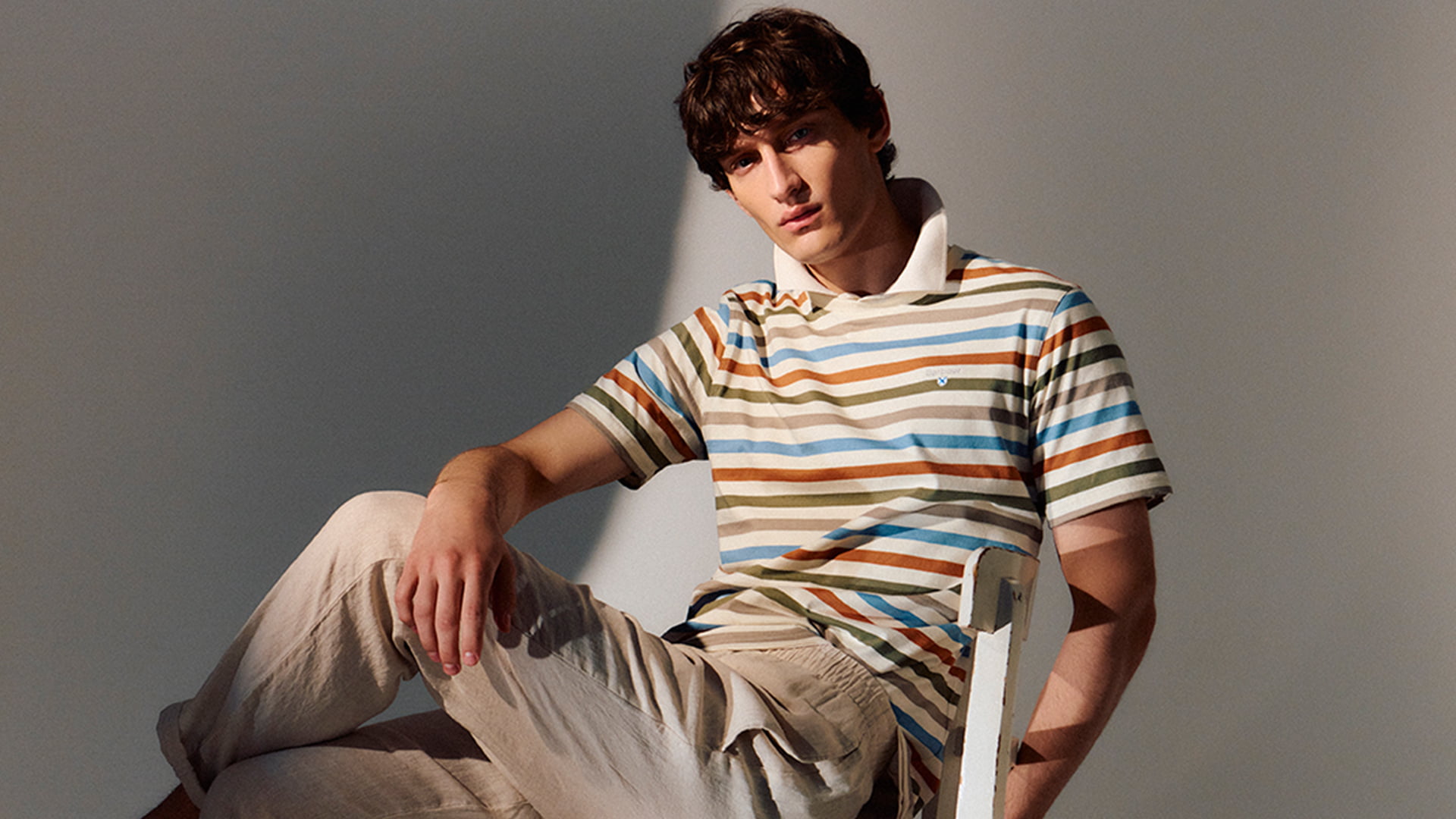 A person in a striped polo shirt and beige pants sits on a white chair, posed against a neutral background.