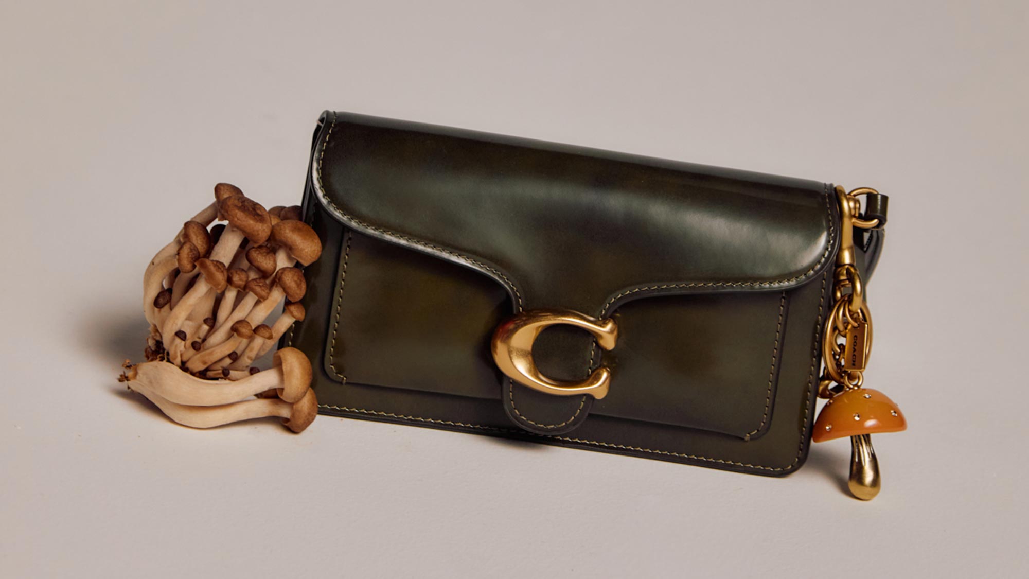 A green leather bag with a gold clasp is adorned with a mushroom charm and surrounded by a small bundle of mushrooms.