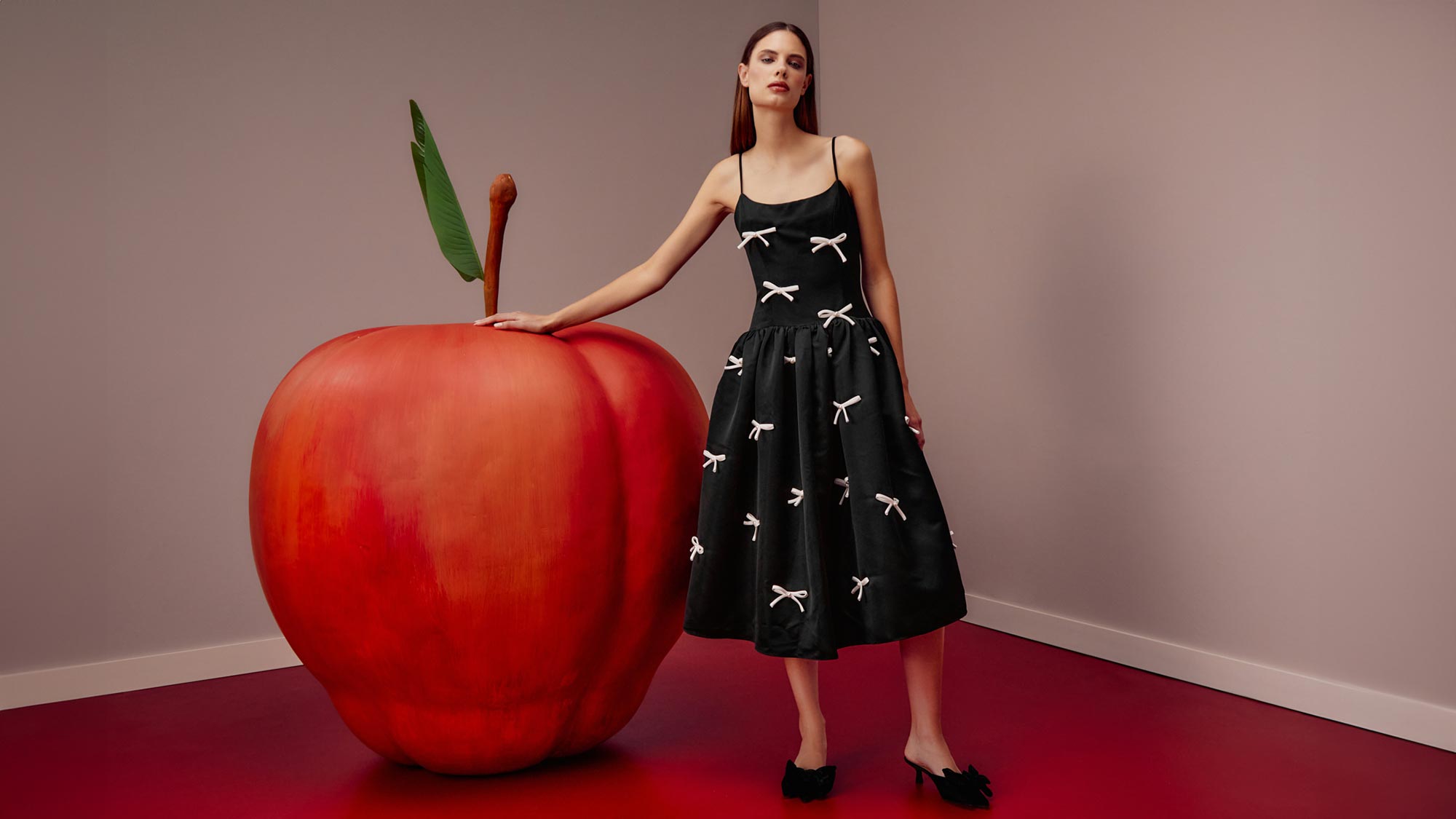 A woman in a black dress with white bows stands beside a giant apple in a minimalist room with red flooring.