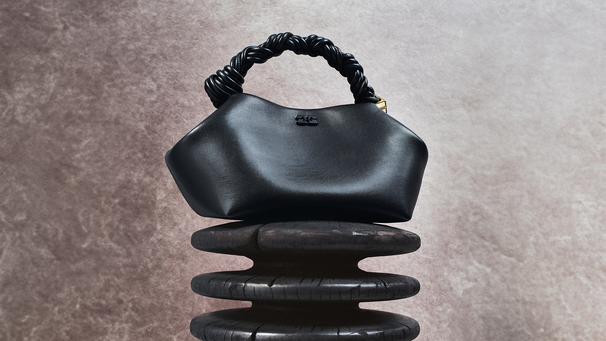 A black Ganni bag with a twisted handle is displayed on a stack of dark, rounded stones.