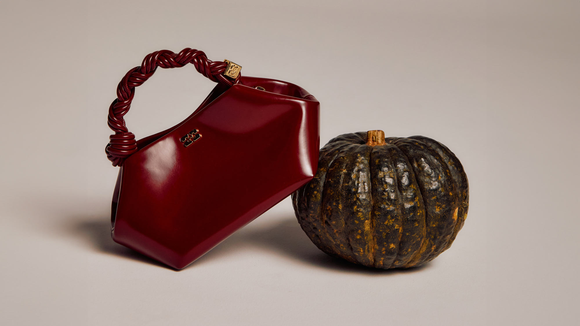 A maroon handbag with a twisted handle is placed next to a round, dark green pumpkin on a plain background.
