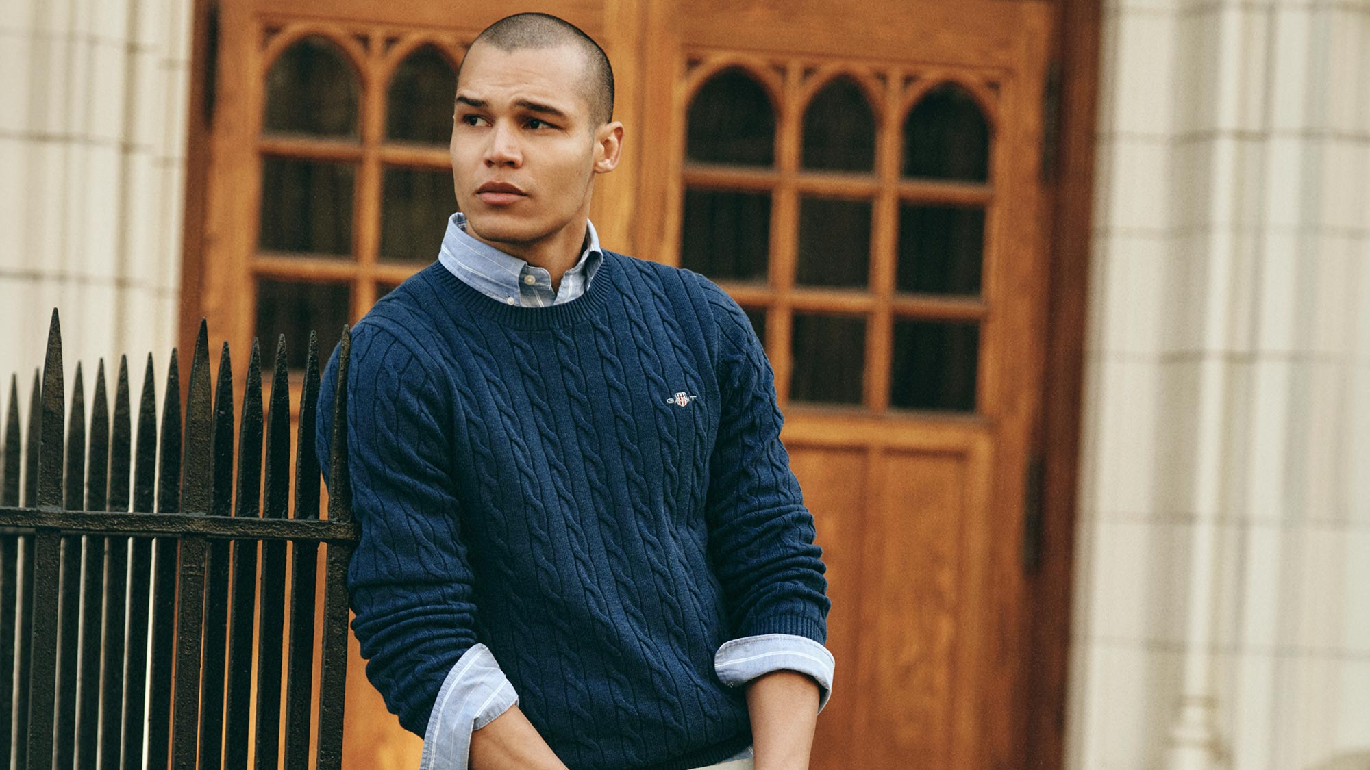 A person in a blue cable-knit sweater and jeans leans on a railing outside a wooden door, holding a book.