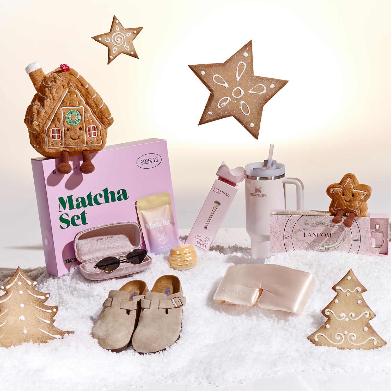 Holiday-themed display with gingerbread cookies, a Matcha set, skincare, sunglasses, a tumbler, slippers, and a pouch on faux snow.