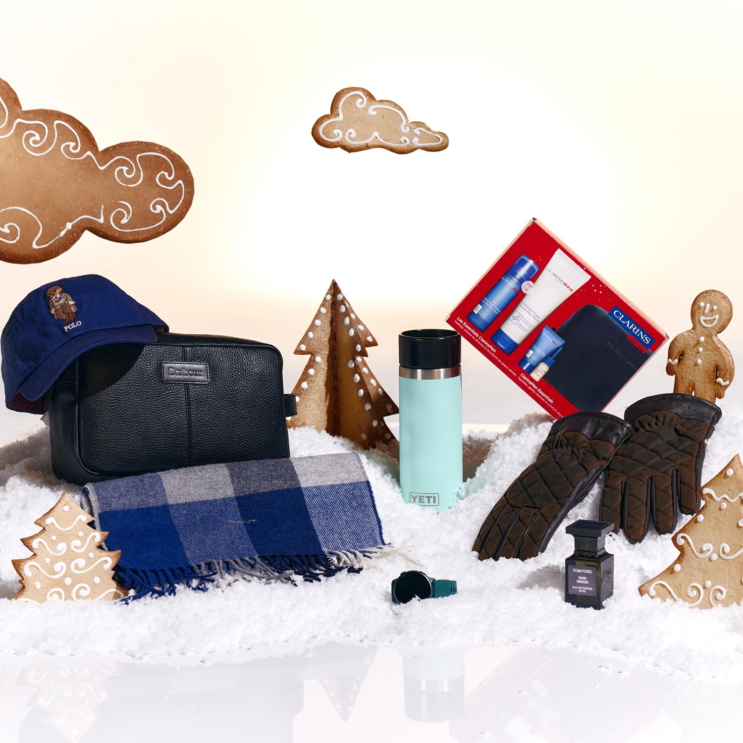 Winter-themed arrangement with gloves, scarf, cap, a thermos, toiletries set, cologne, and gingerbread cookies among fake snow and trees.