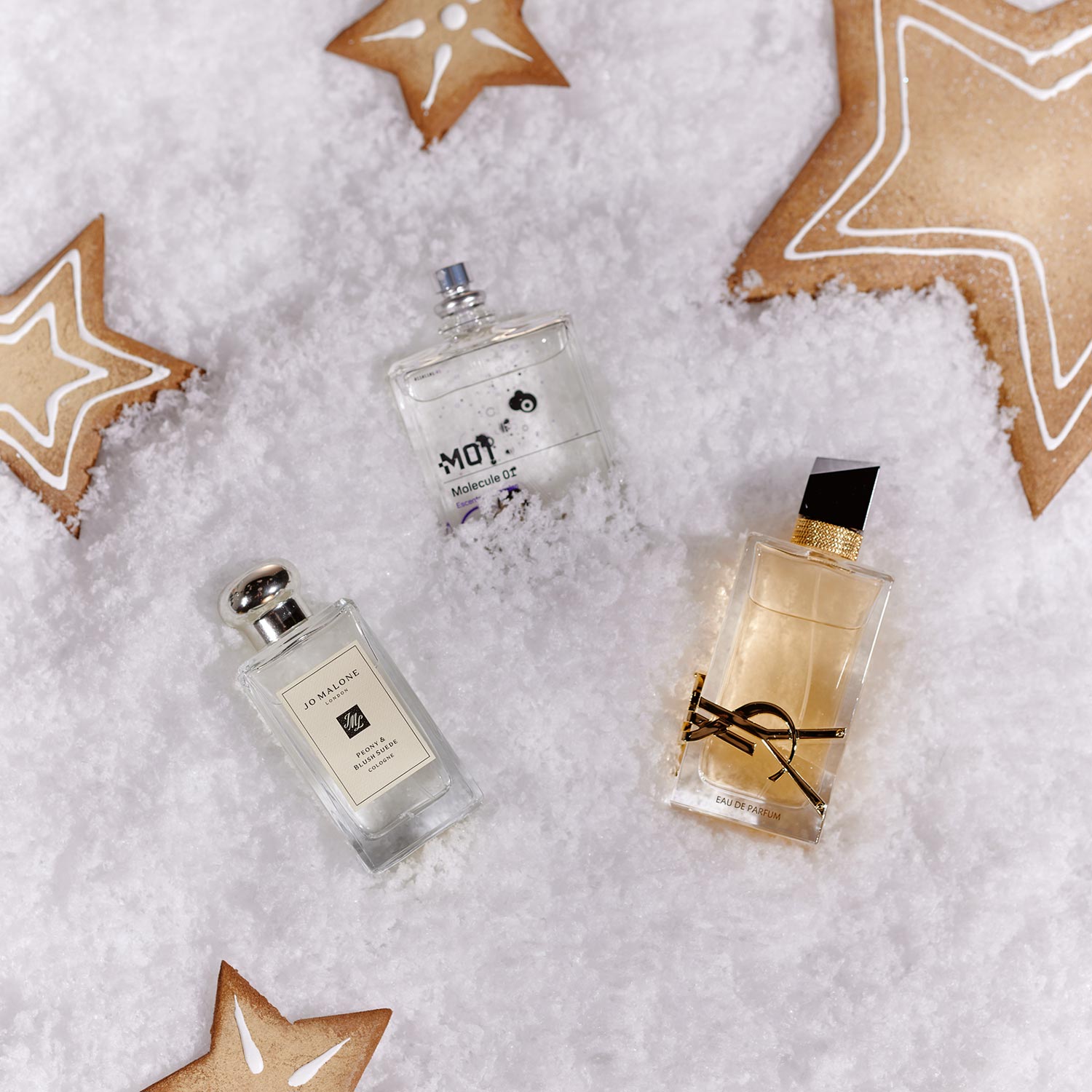 Three perfume bottles are placed on faux snow, surrounded by star-shaped gingerbread cookies.