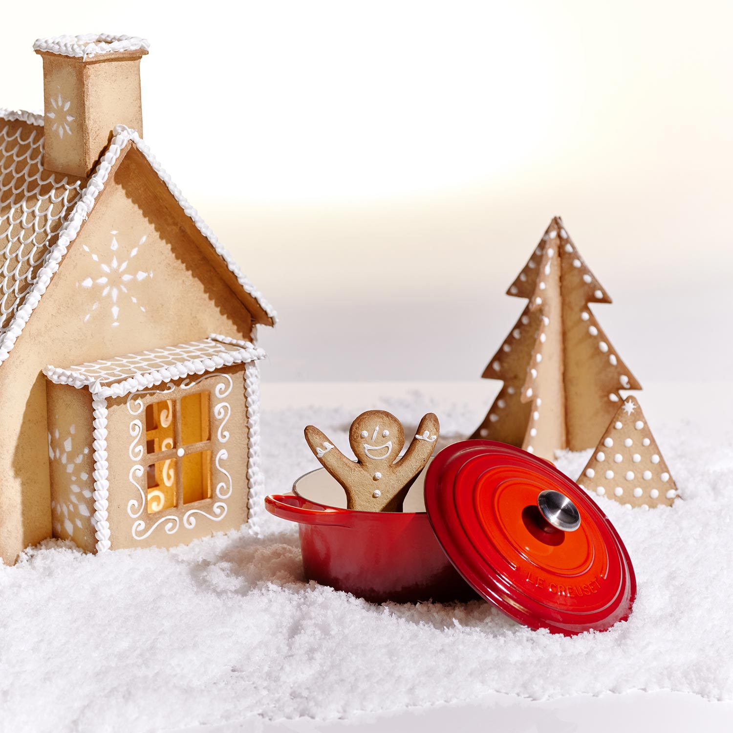 A gingerbread house and trees on snow, with a gingerbread person in a red pot.