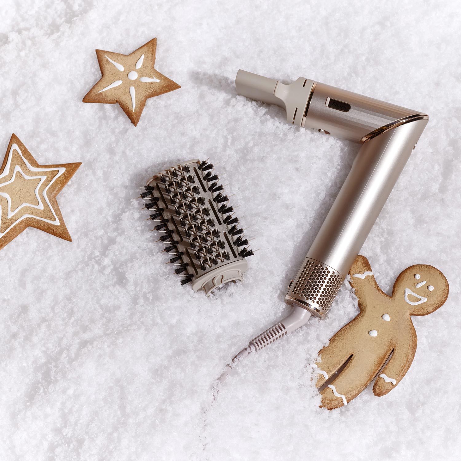 A hair styling tool with a round brush attachment is laid on snow-like powder, surrounded by gingerbread cookies shaped like stars and a person.