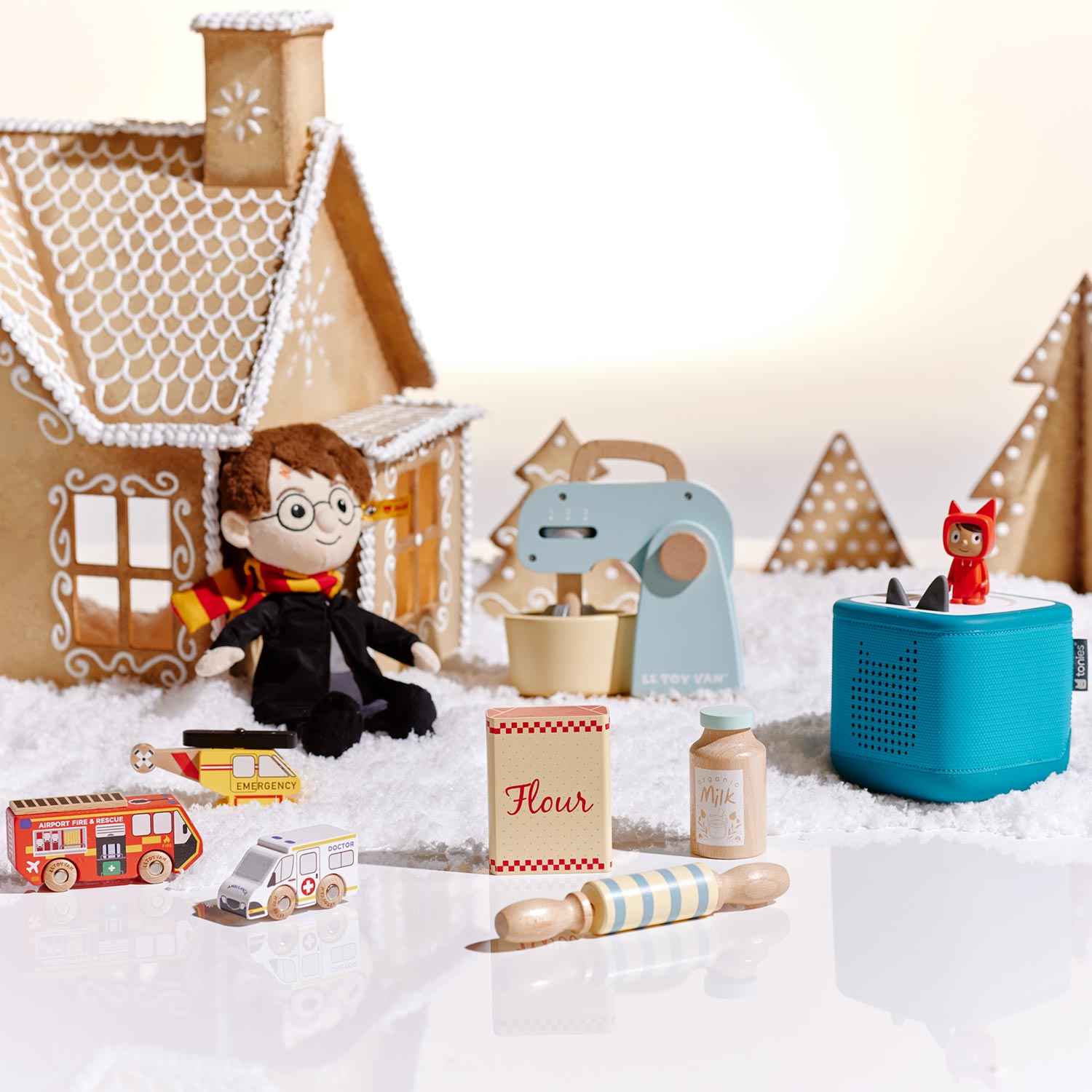 A gingerbread house with a plush toy and miniature toys, including a mixer, flour container, toy vehicles, and a blue speaker, in a snowy setting.