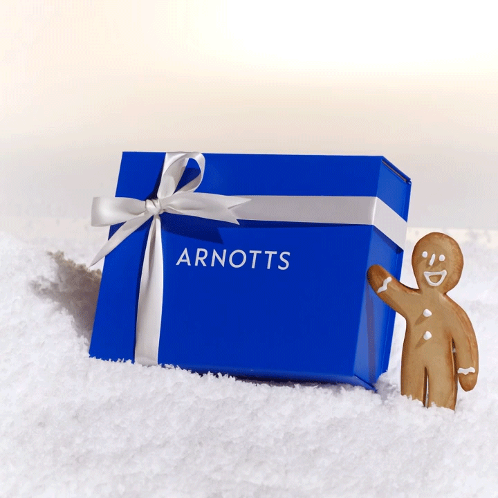 A blue gift box with a white ribbon labeled 'ARNOTTS' sits in the snow next to a smiling gingerbread figure.