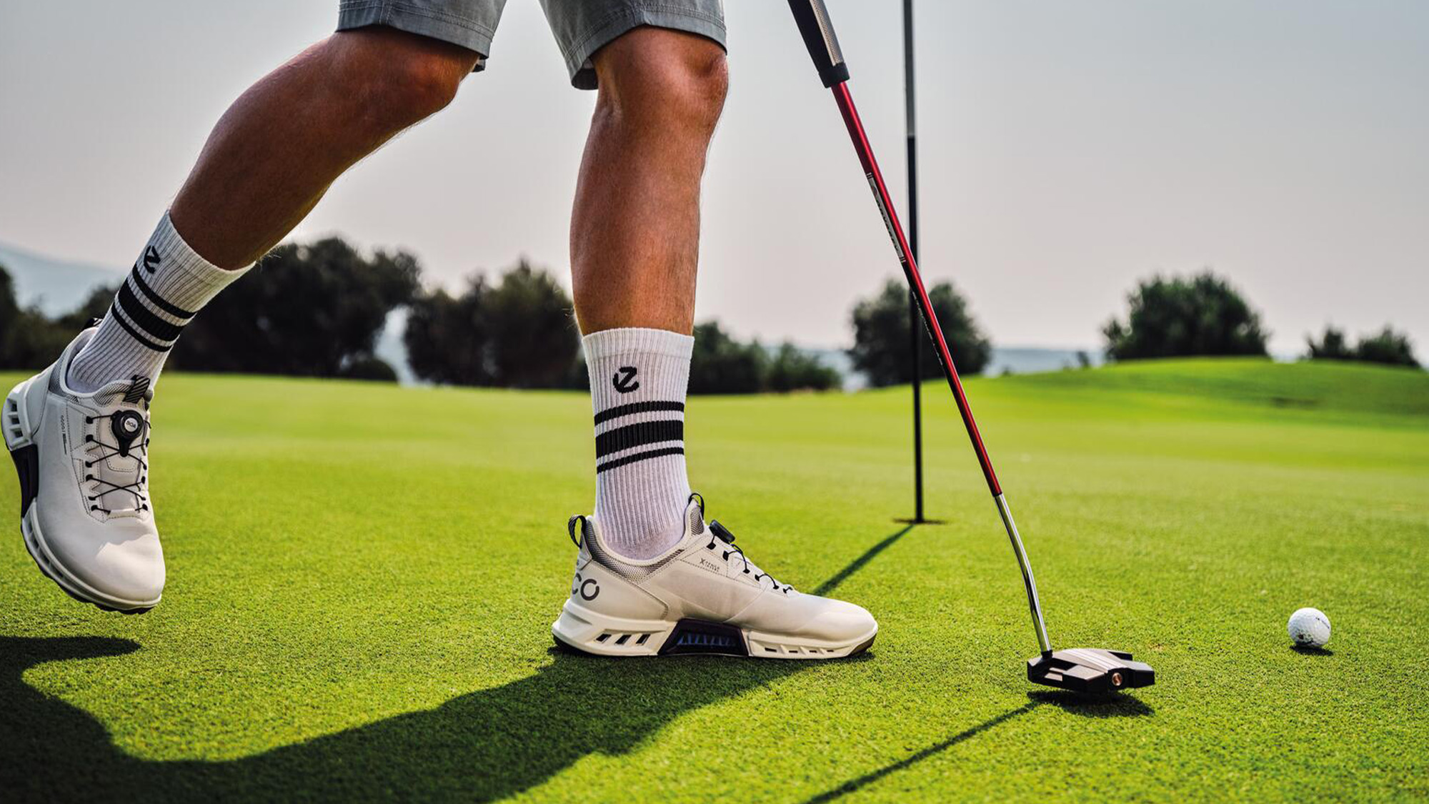 Golfer in shorts and white shoes putting on a green with a ball and flag nearby.
