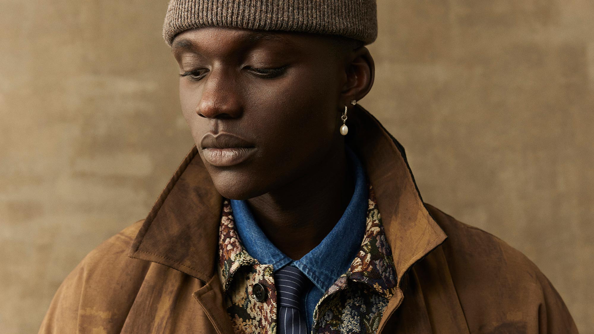 A person in a brown beanie and coat wears a floral shirt, tie, and earring, standing against a plain background.